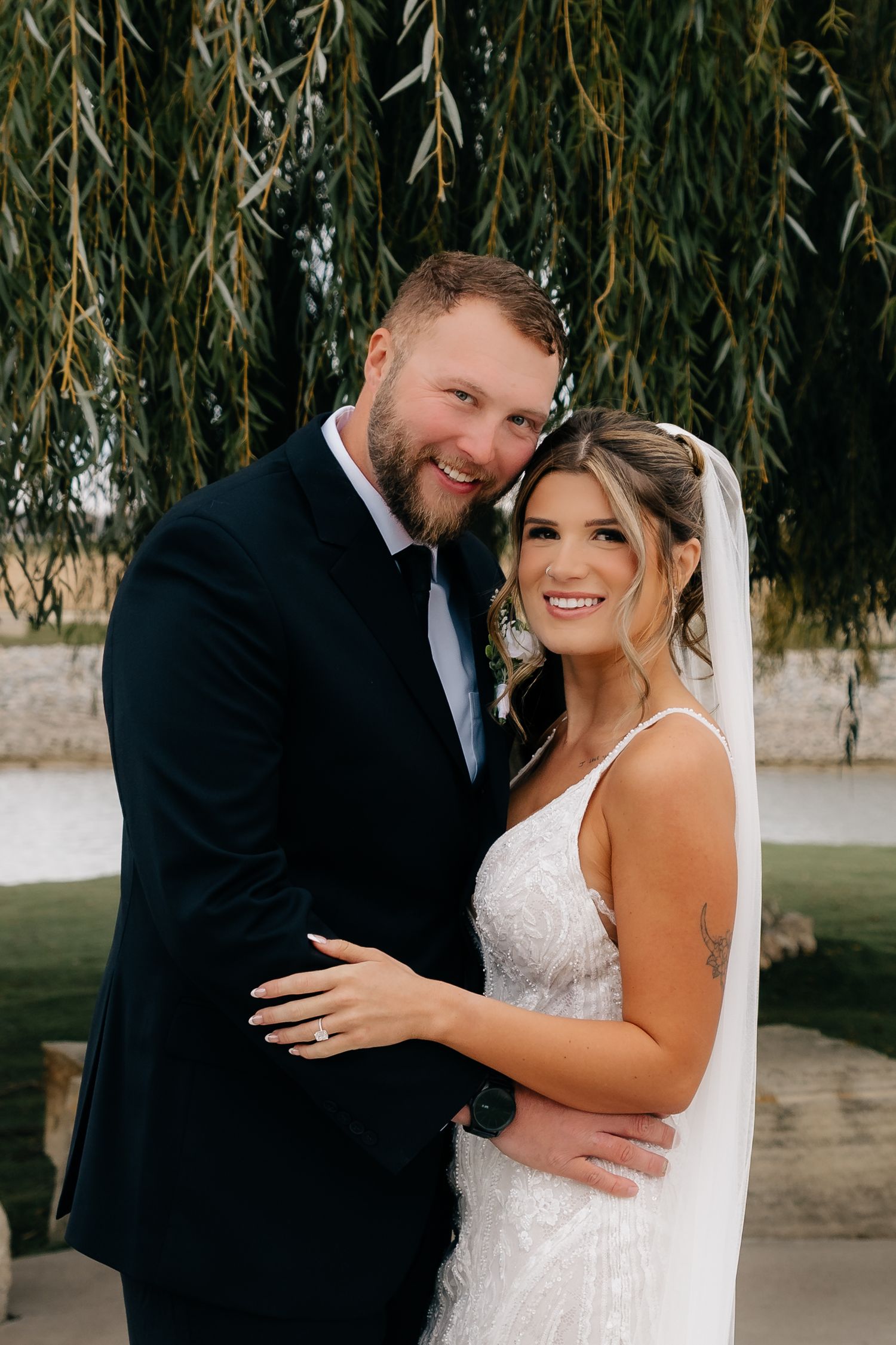 Bride and groom smiling together under weeping willow tree on wedding day.