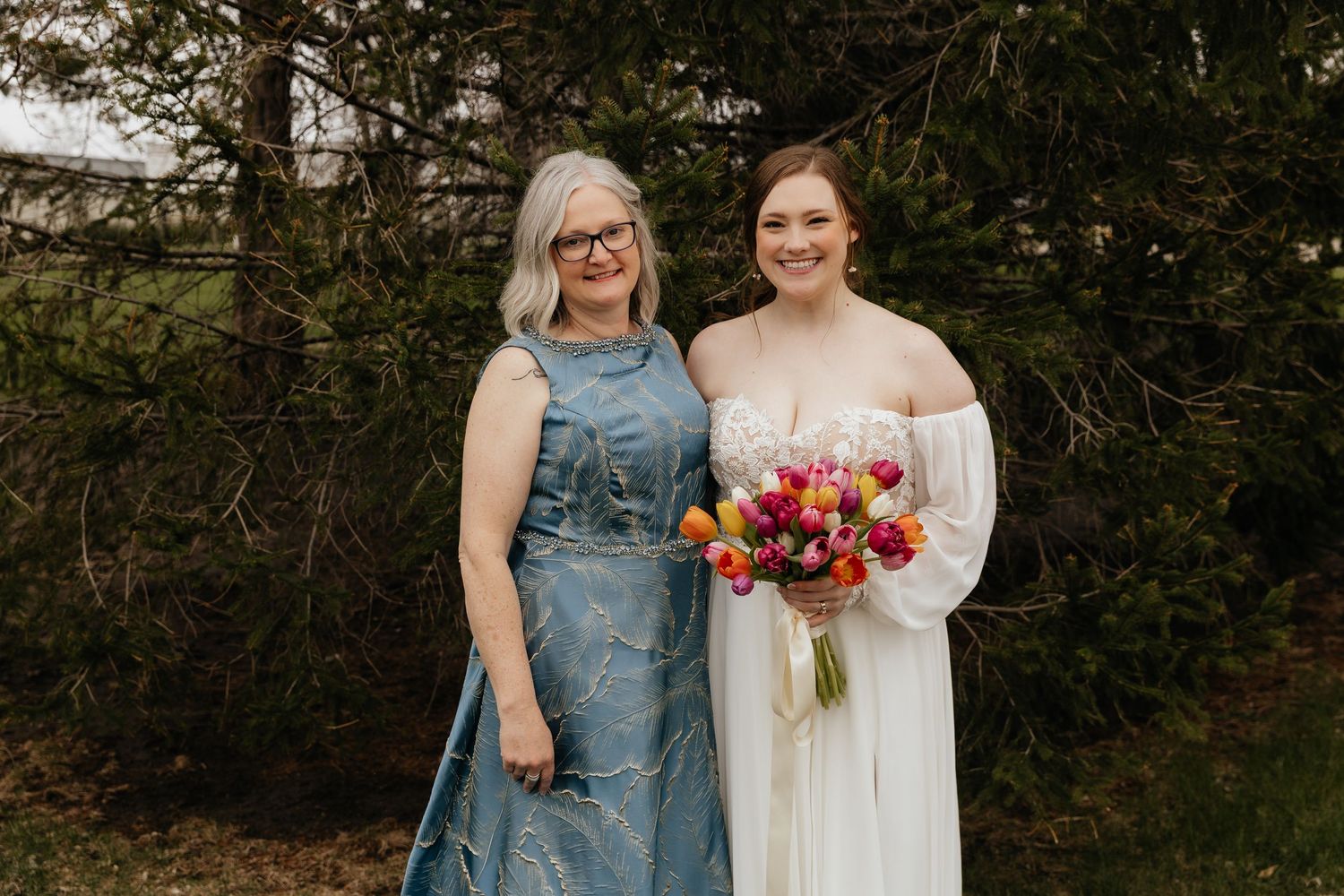 Bride holding vibrant tulip bouquet smiles alongside mother in blue gown outdoors.