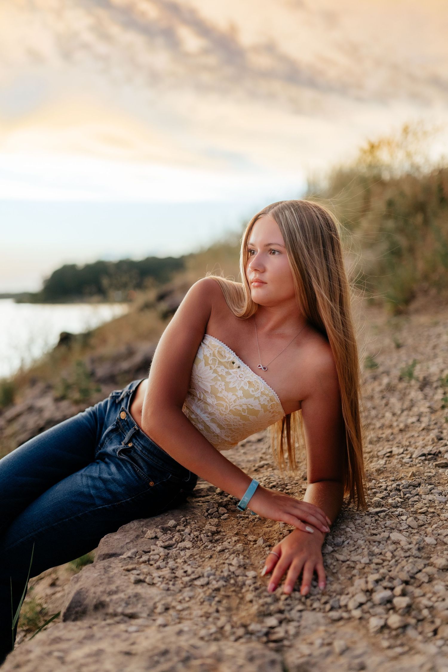 Blonde teenage girl in tube top and jeans reclines on rocky ground near water at golden hour.
