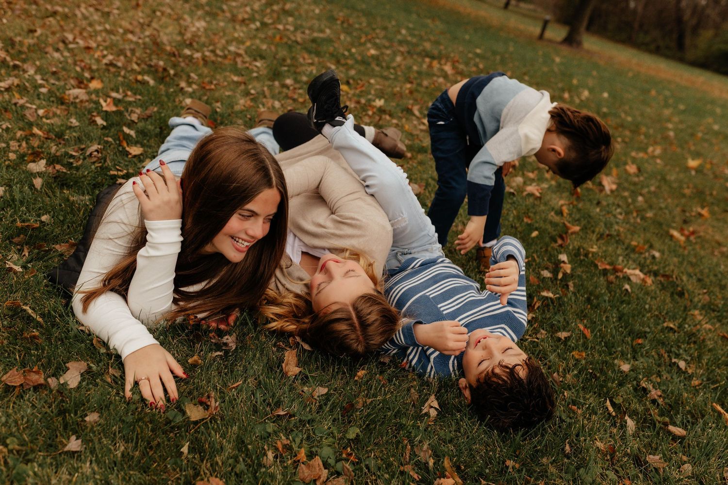 Four siblings laughing and laying together on grass covered with fallen autumn leaves in a park.