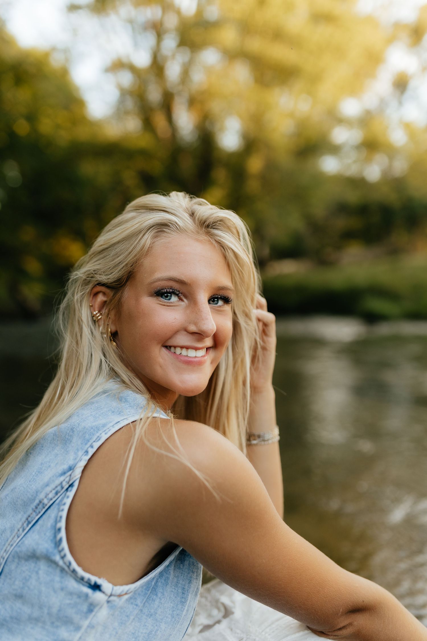 Smiling blonde teen girl near river in denim vest with autumn trees behind her.