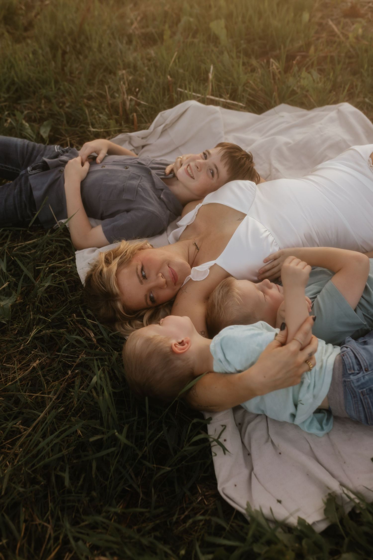 Mother lying in green grass with three children cuddling together during golden hour outdoor session.