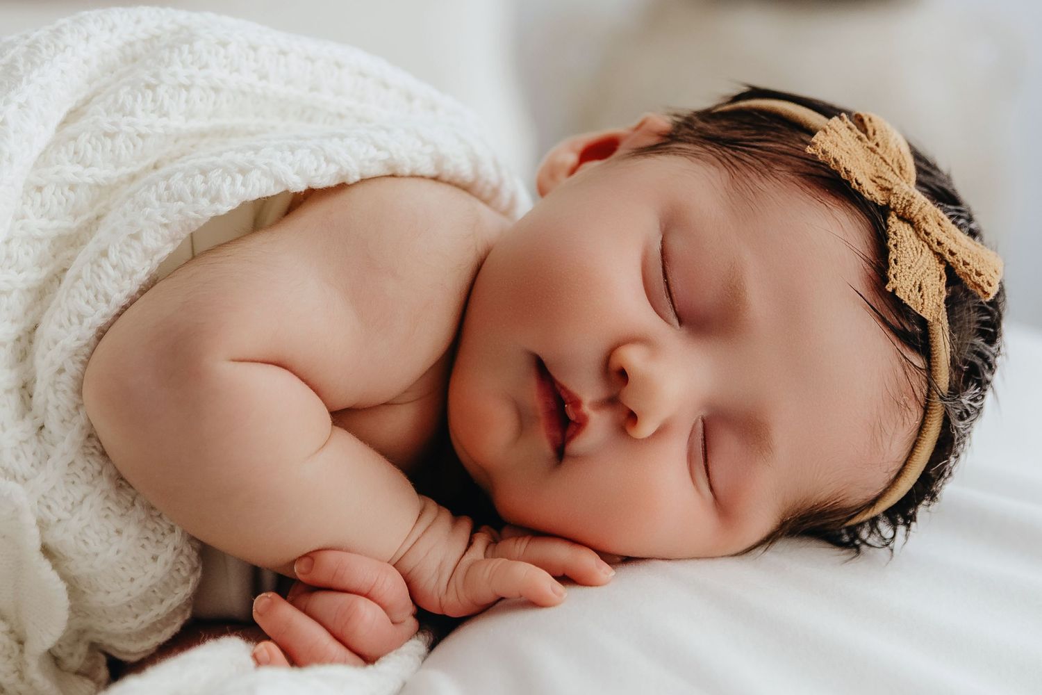 Peaceful sleeping newborn baby with dark hair wearing gold bow headband wrapped in white blanket.