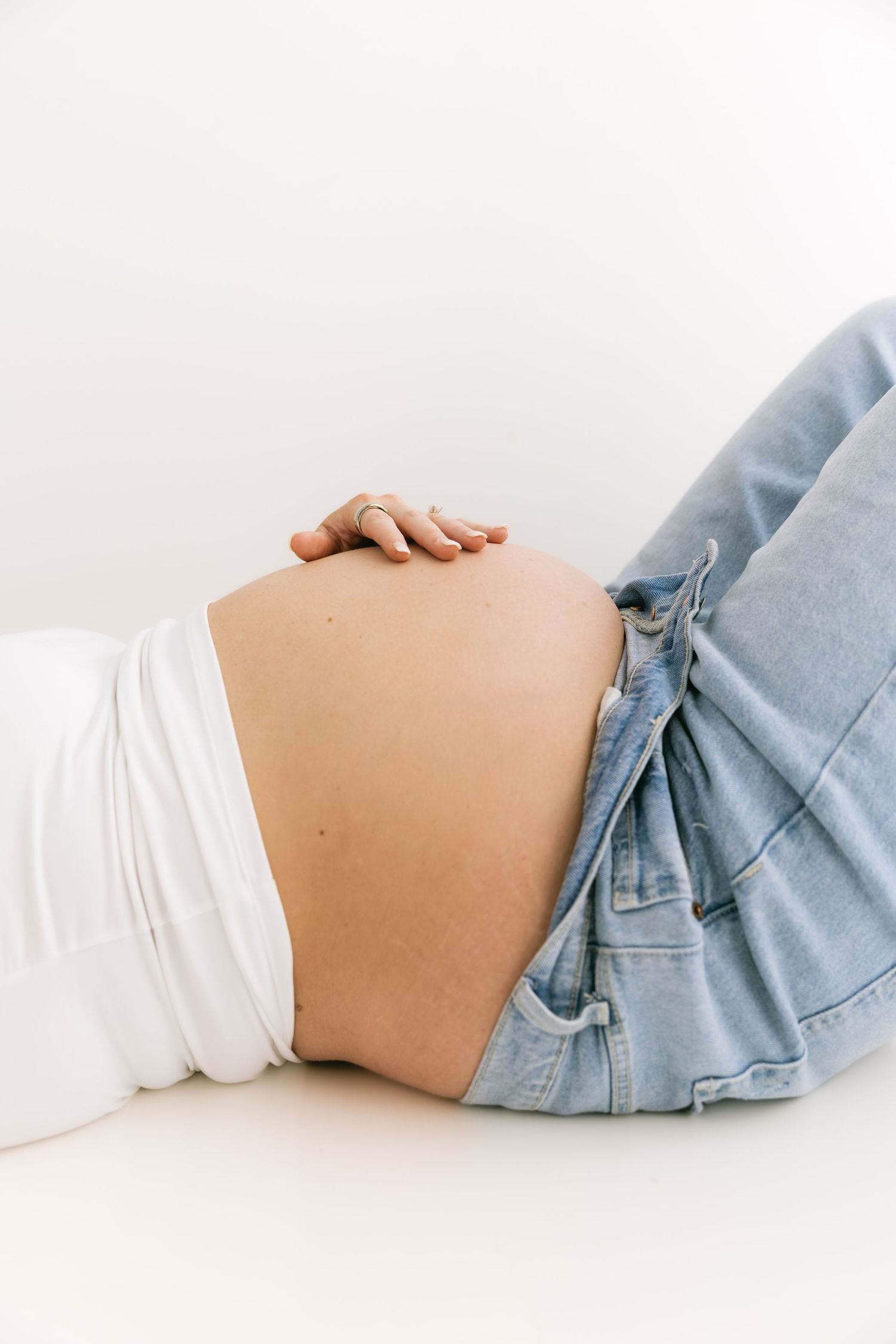 Close-up of pregnant woman lying down, cradling bare baby bump, wearing white top and open denim jeans.