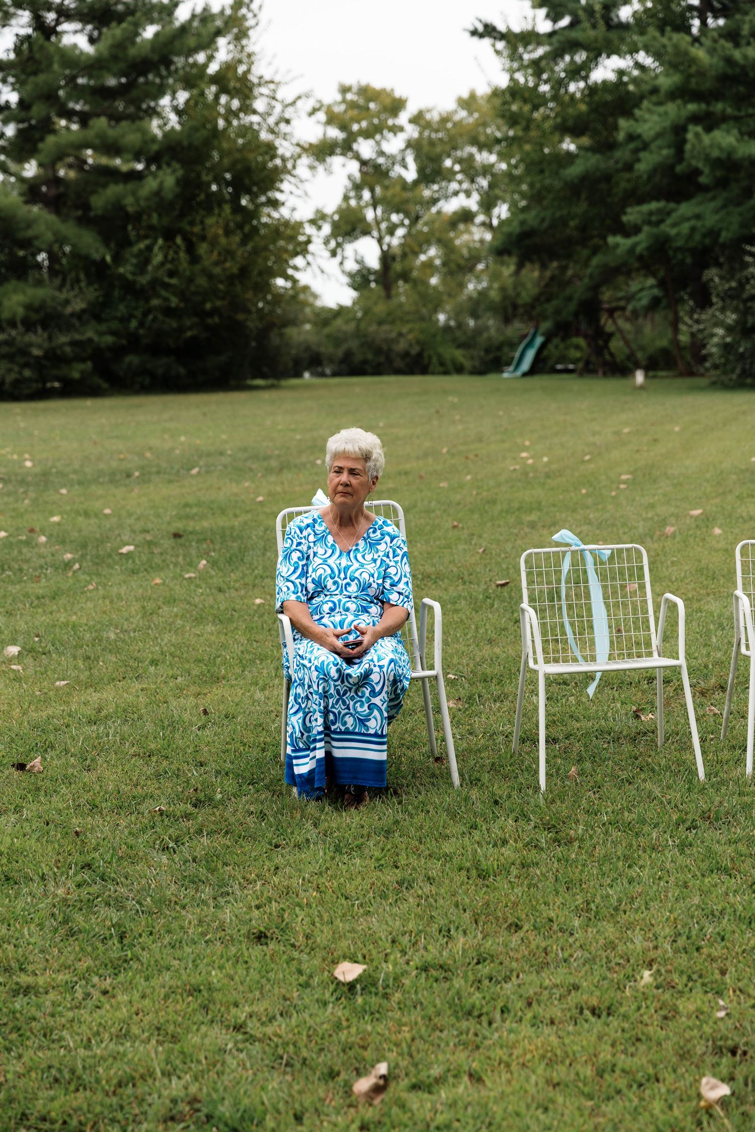 Elderly woman in blue floral dress standing on green lawn near white chairs at outdoor wedding.