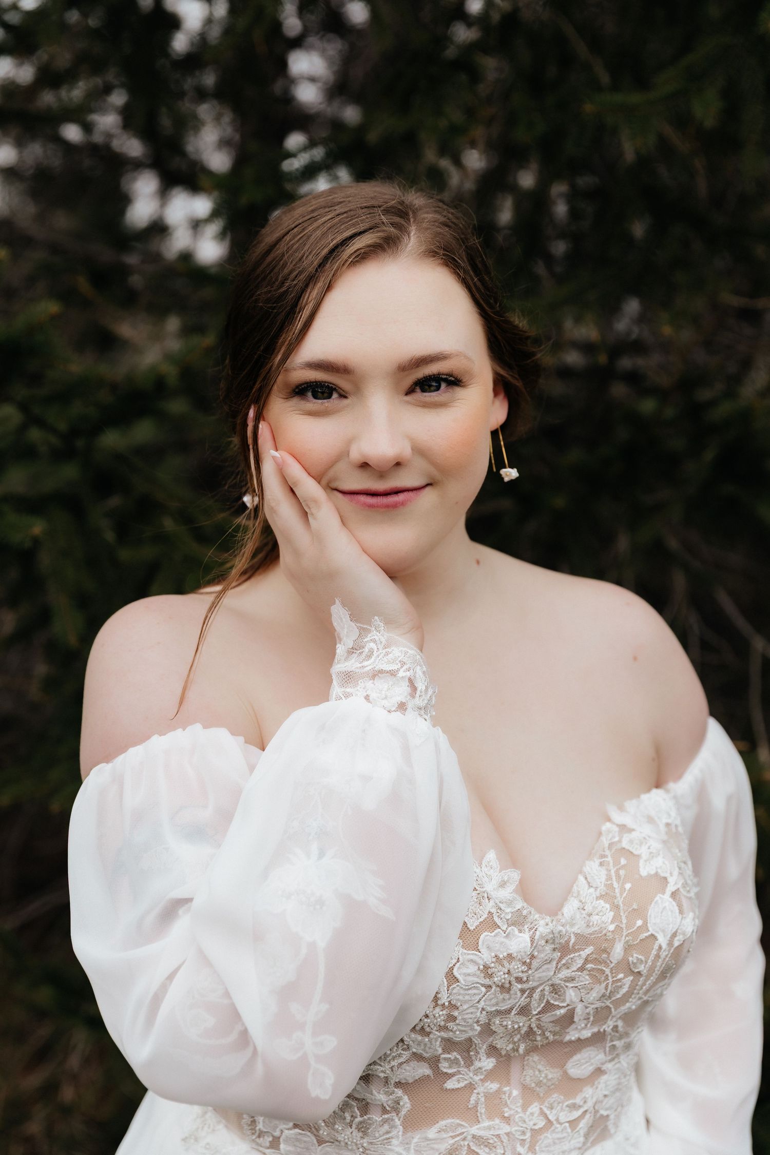 Bride in off-shoulder lace wedding gown poses gracefully with hand on cheek outdoors.