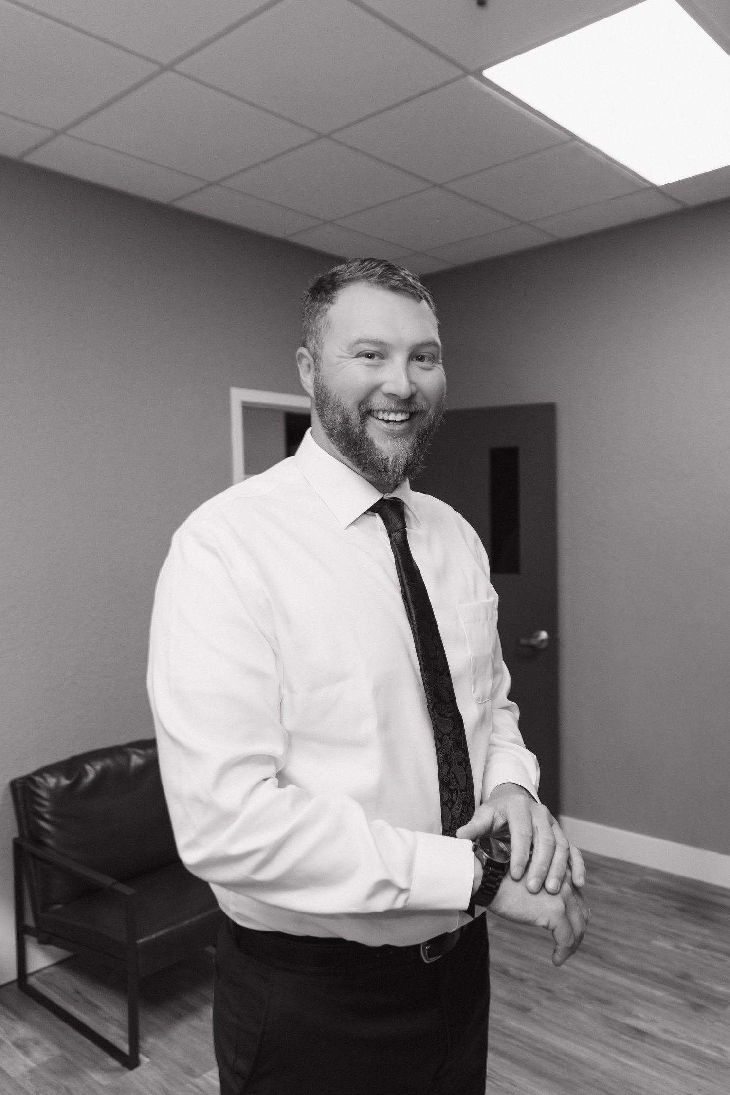 Black and white portrait of a smiling bearded groom in a white shirt and tie checking his watch.