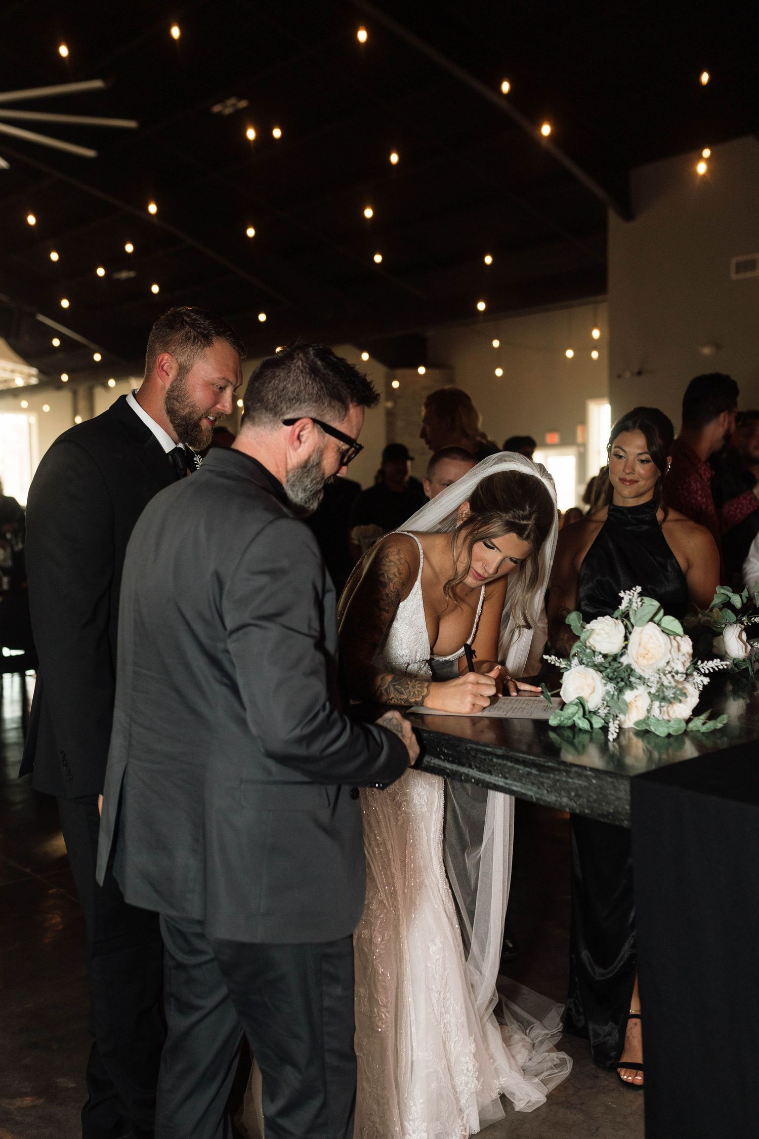 Bride signs marriage certificate at wedding reception surrounded by guests and white floral arrangement.