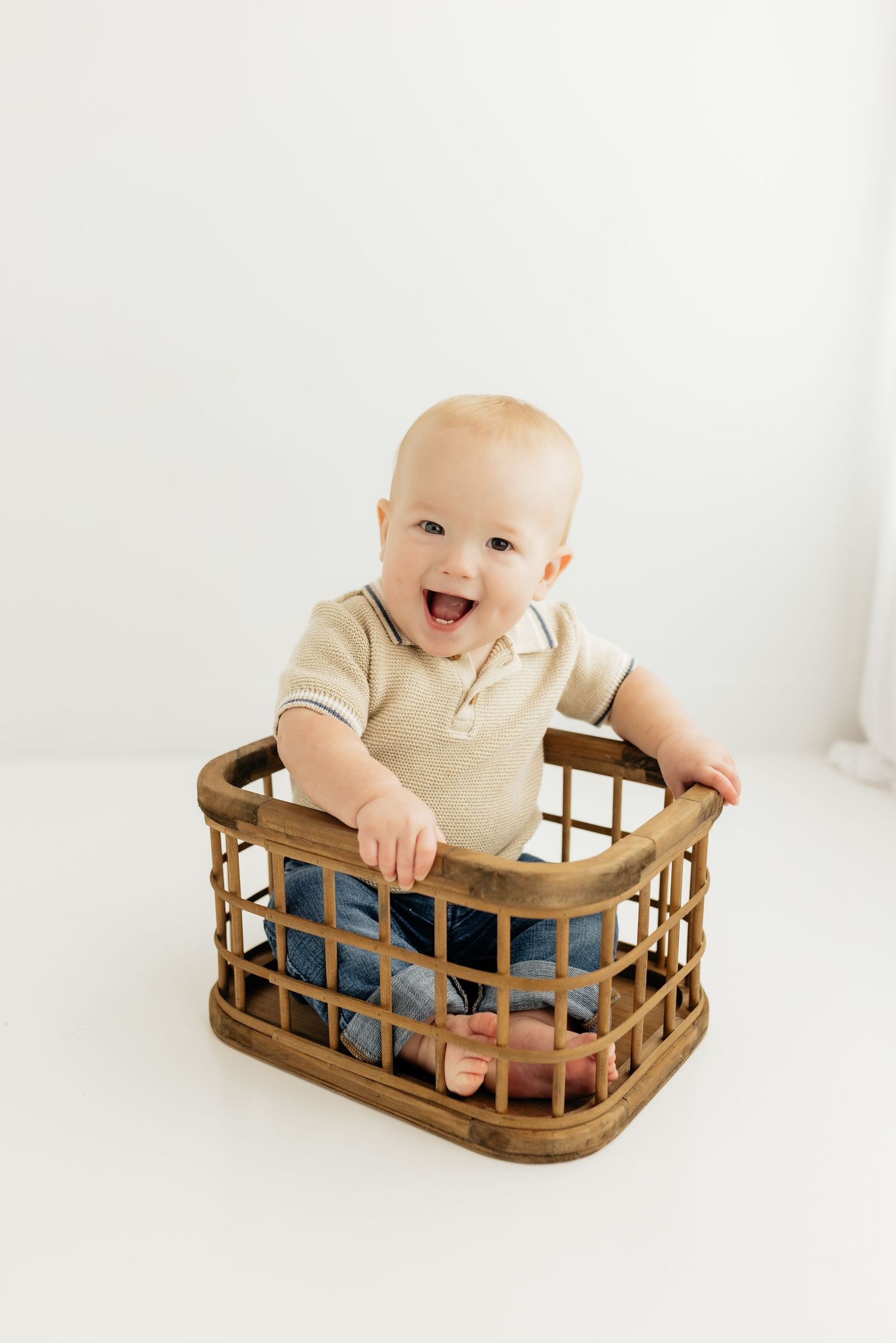 Happy baby sitting in a wooden crate basket, wearing a beige polo shirt and jeans, smiling joyfully.