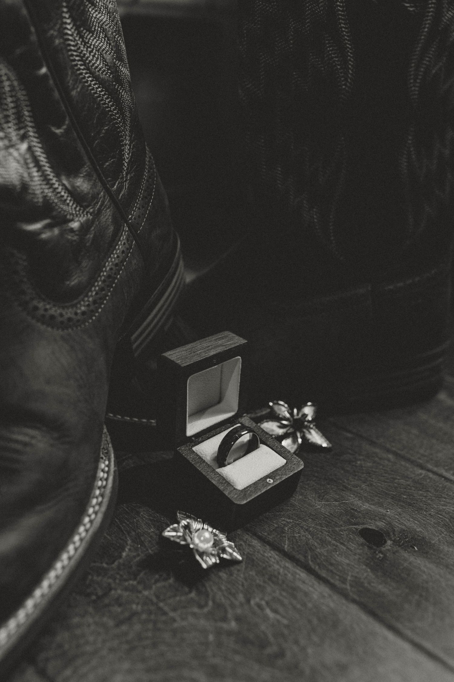Black and white image of cowboy boots beside a ring box with wedding bands and floral accessories.