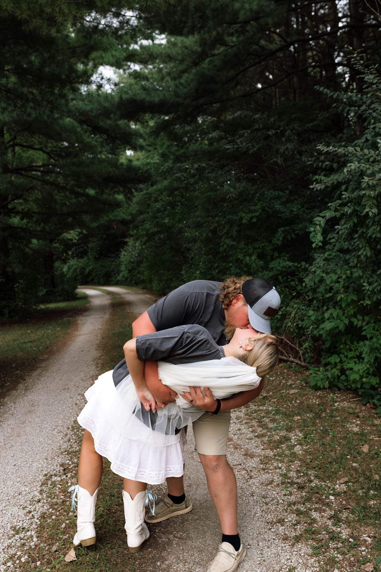 Groom dipping and kissing his bride on a rustic gravel path surrounded by tall pine trees.