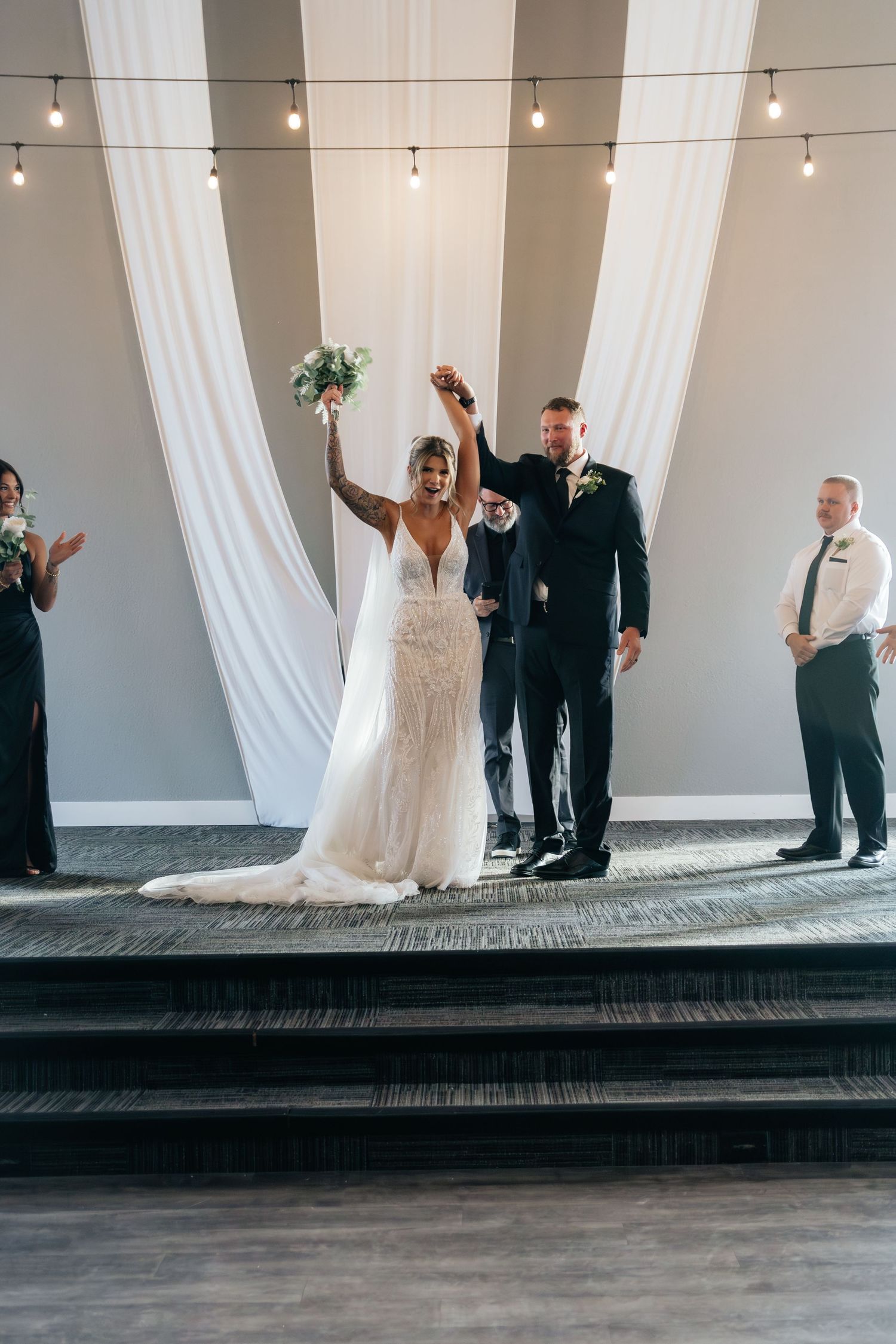 Newlyweds celebrate with raised hands at ceremony altar with white draping and string lights.
