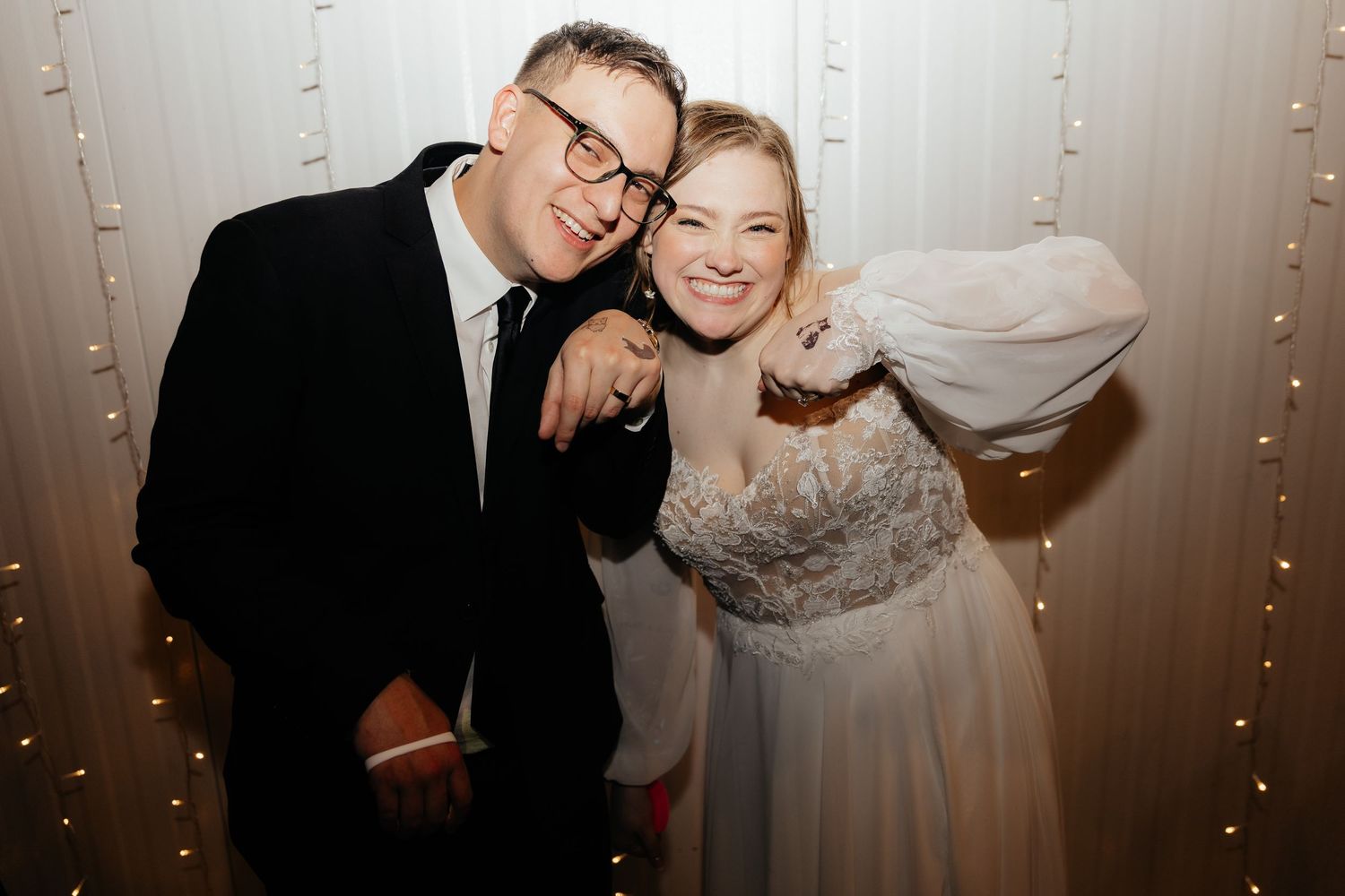 Laughing bride and groom pose together in front of glowing white fairy light backdrop at wedding reception.