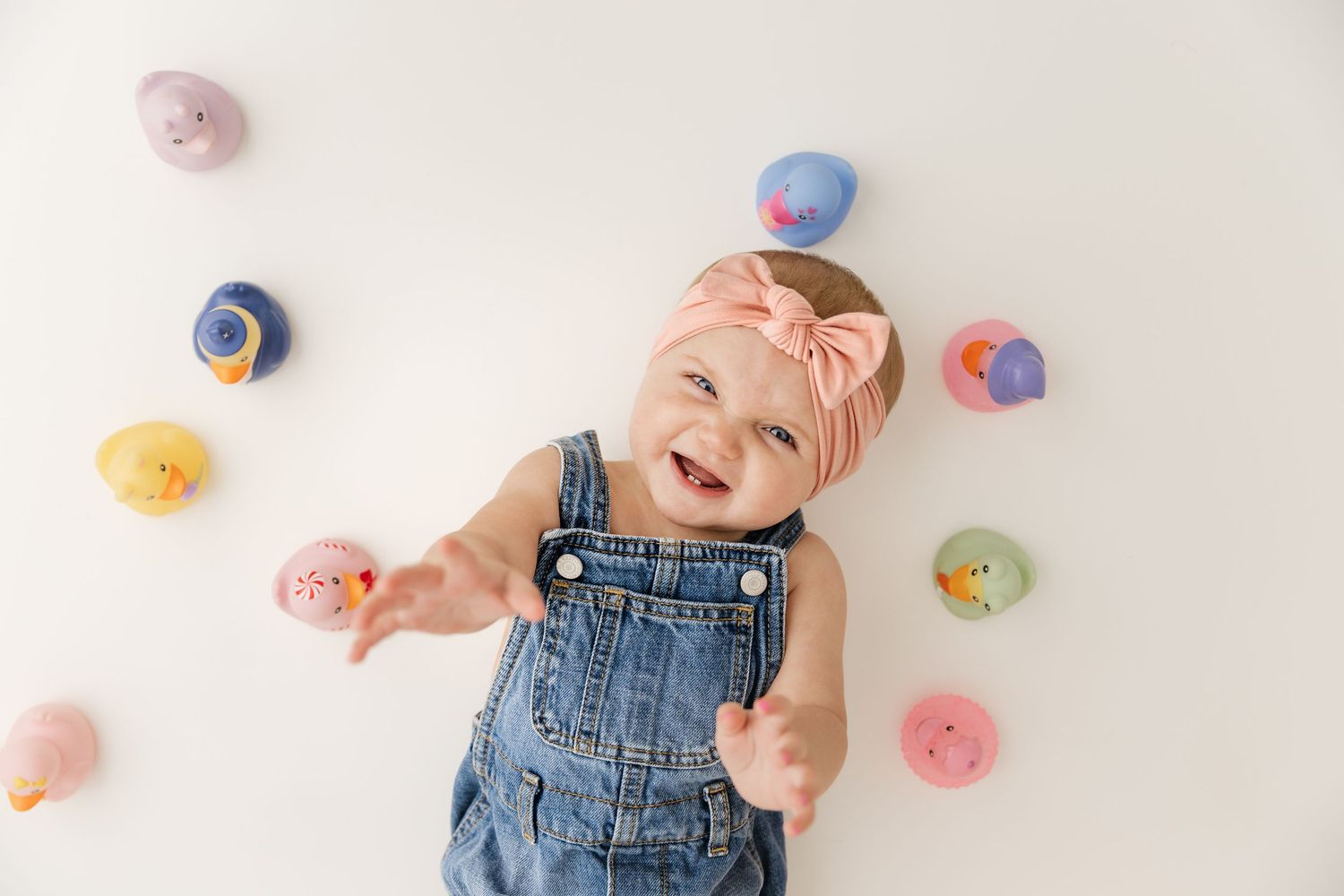 Happy baby girl in denim overalls and pink bow headband laughing while lying among colorful rubber ducks.