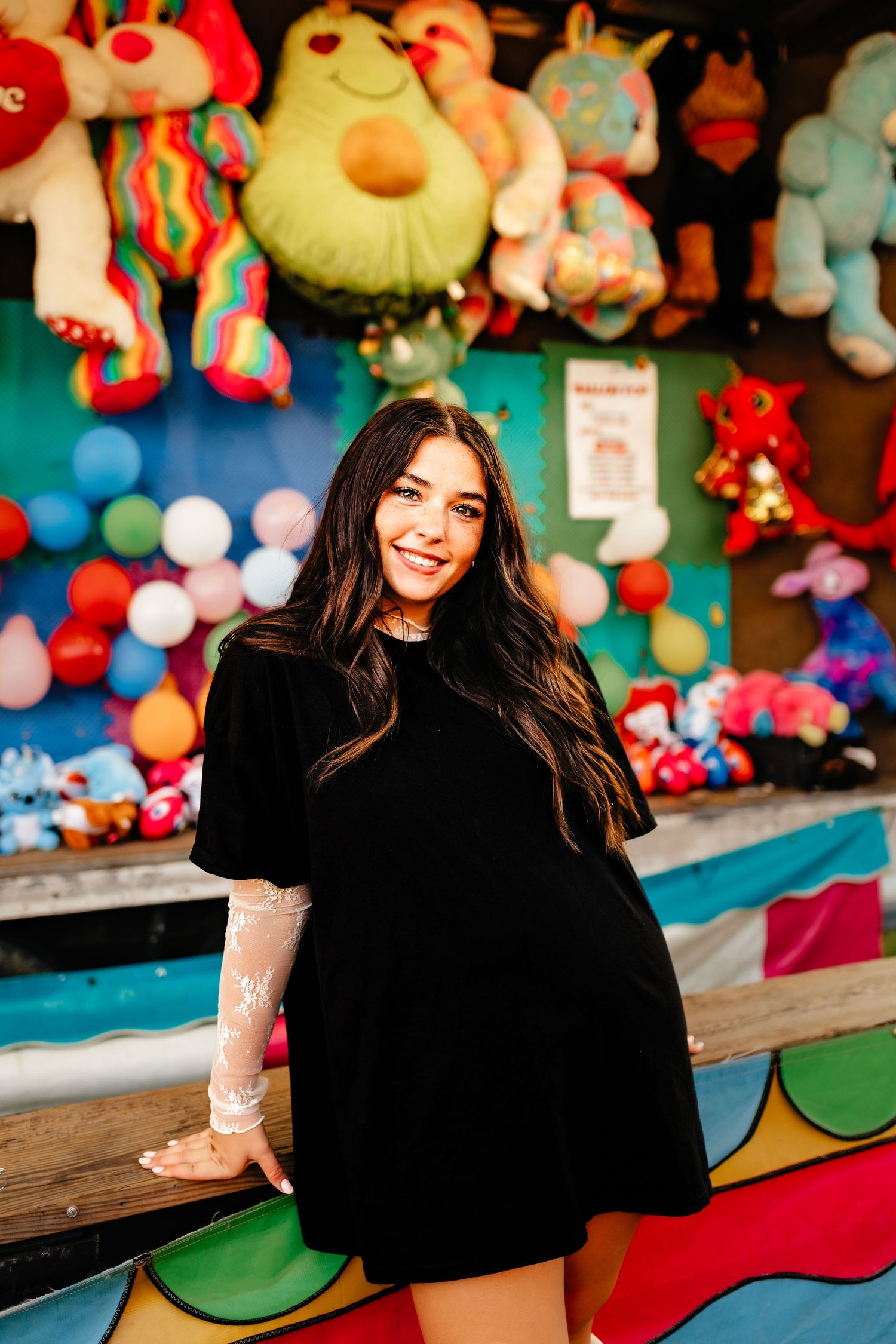 Smiling young woman in black dress posing at colorful carnival game booth with stuffed animals.
