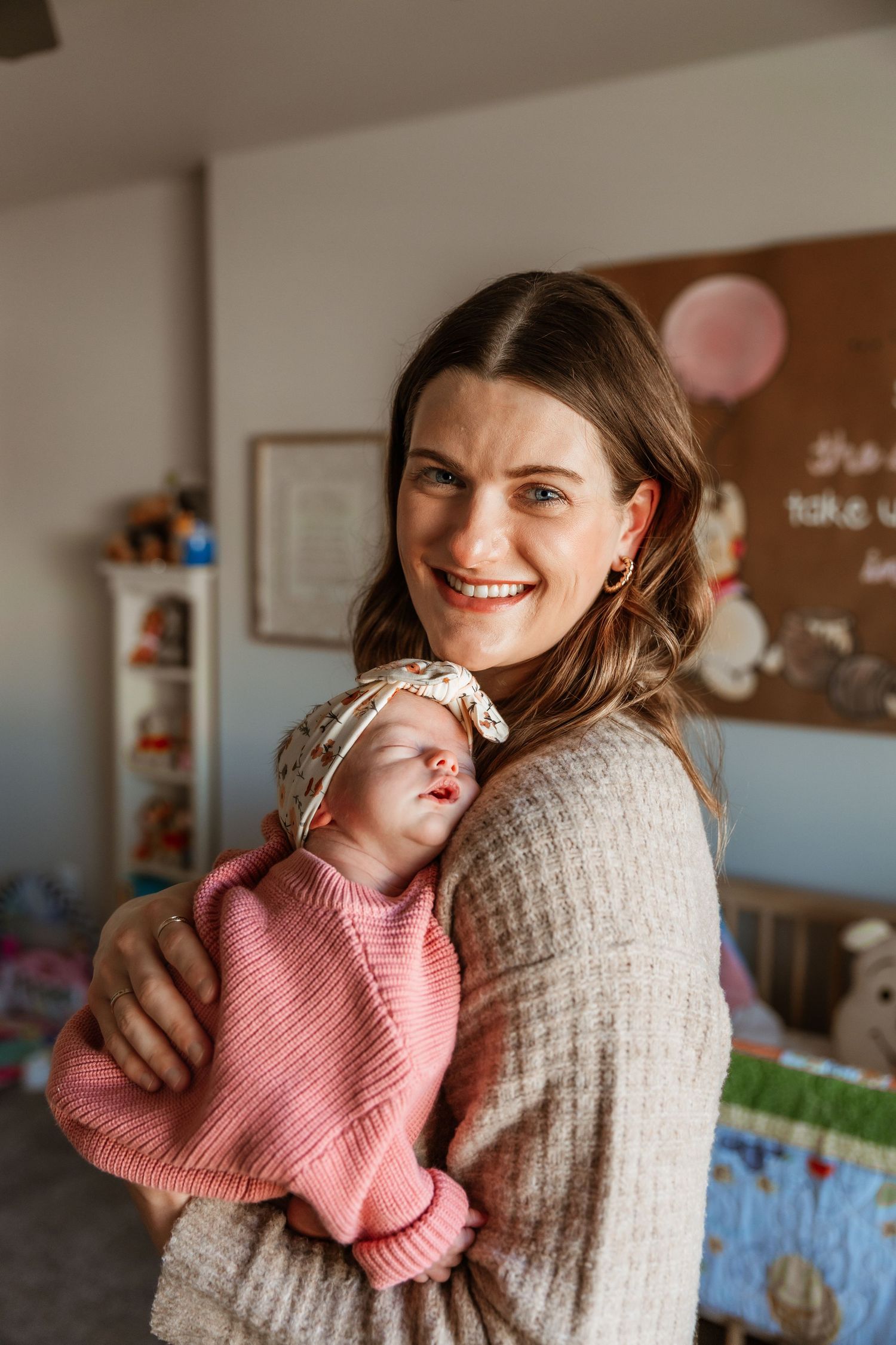 Happy mother in beige sweater holds her sleeping newborn baby girl in pink in nursery.