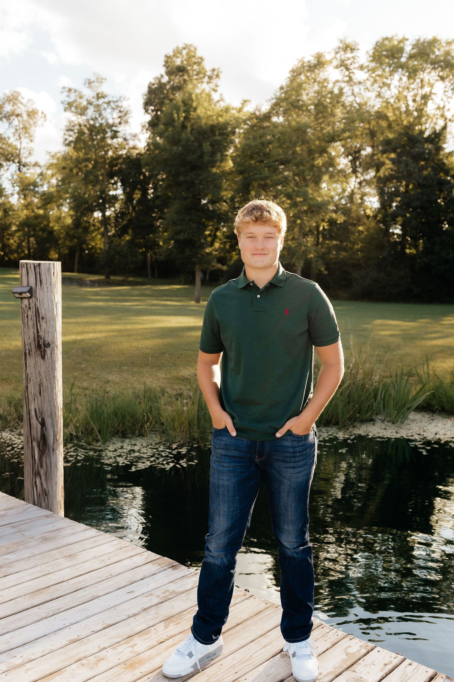 Young man in green polo shirt posing on a wooden dock by a serene pond at sunset.