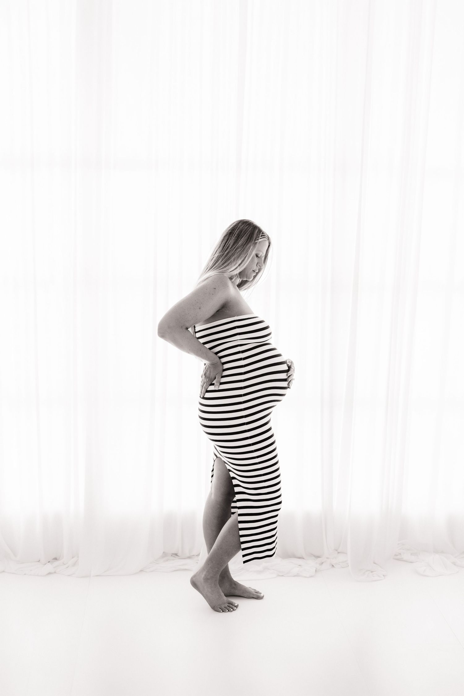 Black and white profile of pregnant woman in striped strapless dress, hands on belly, standing by sheer curtains.