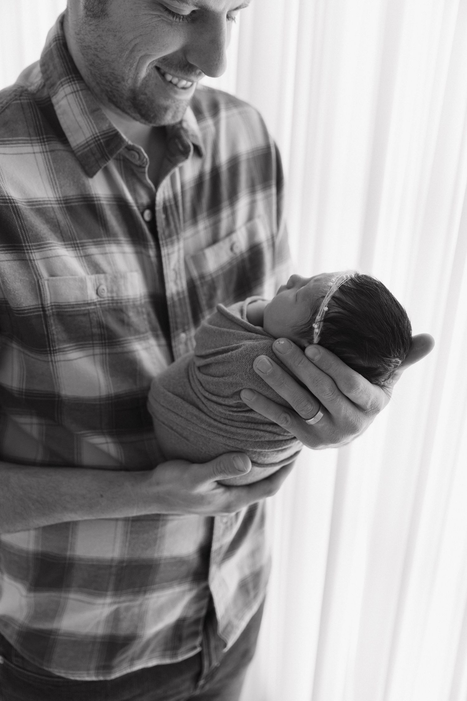 Black and white photo of smiling father in plaid shirt gently cradling tiny sleeping newborn.
