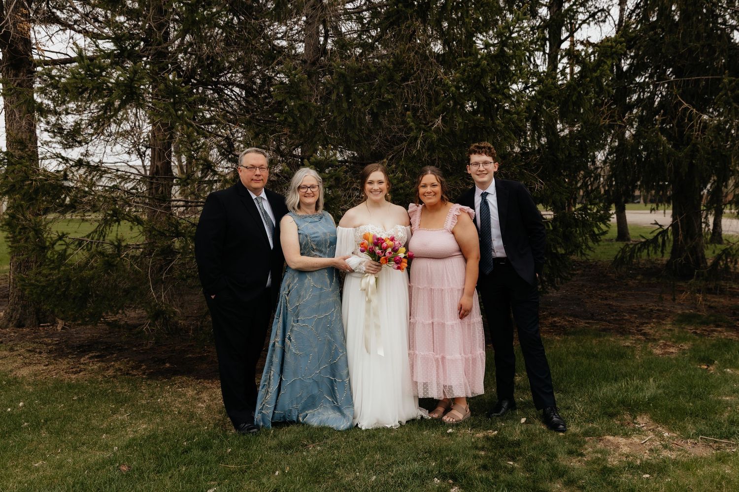 Bride in white gown poses with four family members for outdoor wedding portrait near trees.