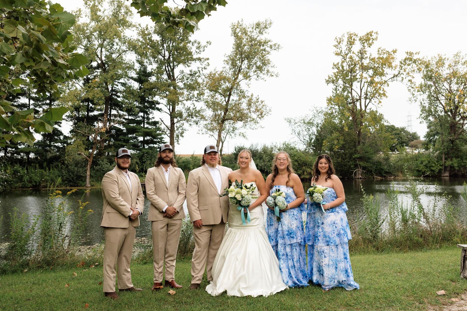 Wedding party poses by pond with bride, groom, groomsmen in tan suits and bridesmaids in blue.