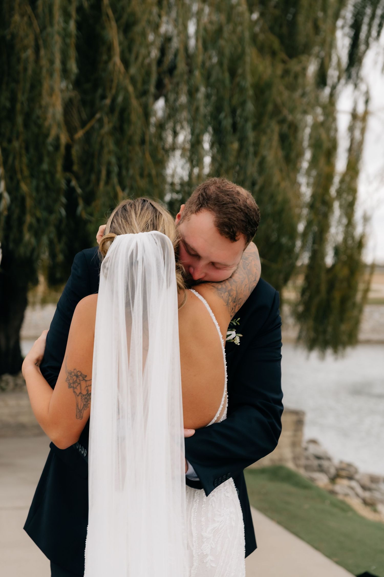 Bride and groom sharing emotional first look embrace under weeping willow tree by waterfront.