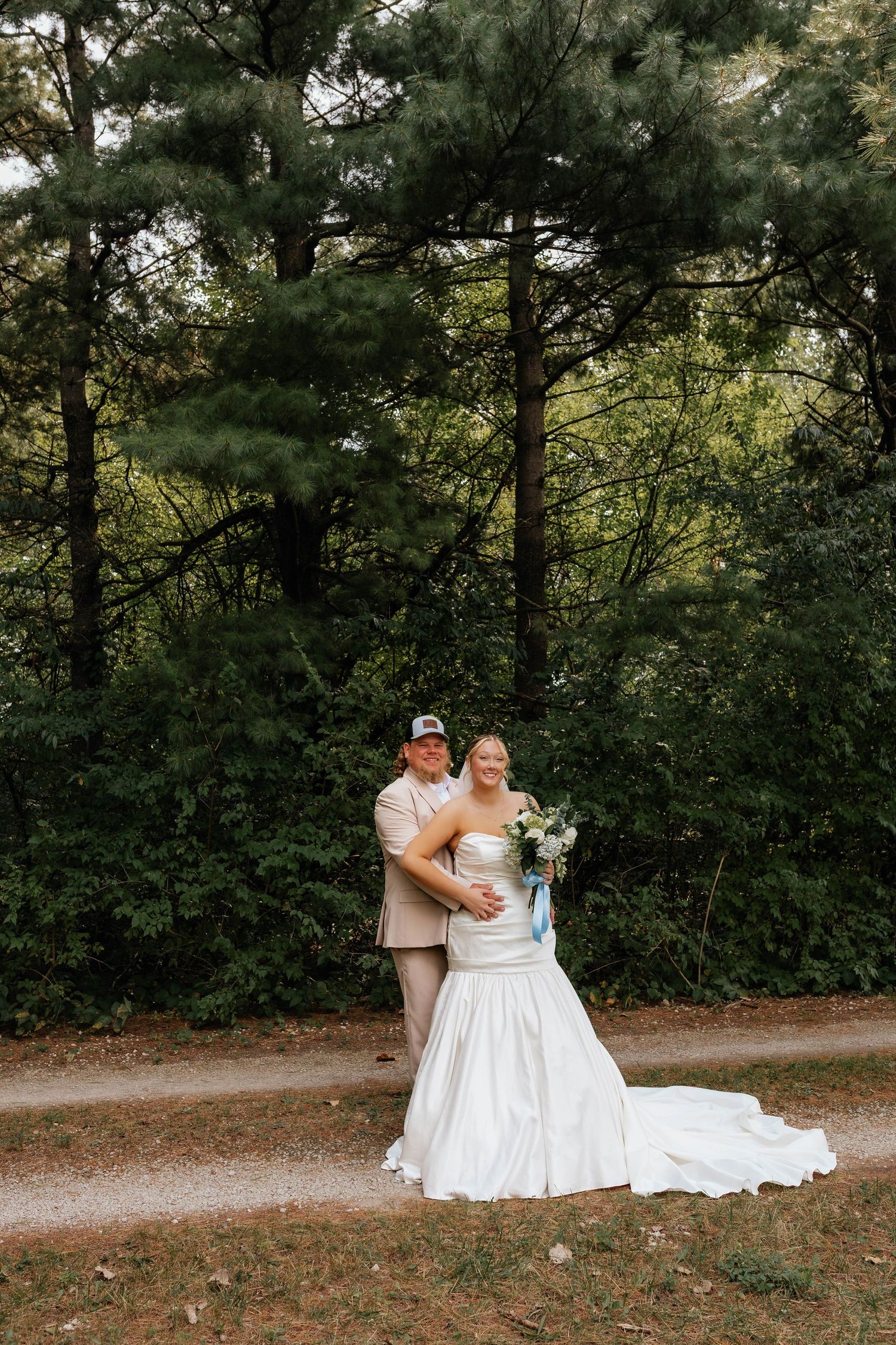 Wedding couple poses on forest path, groom in tan suit embracing bride in white gown with bouquet.