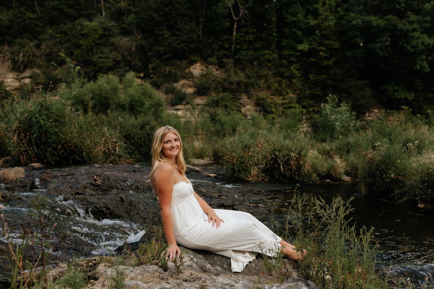 Blonde woman in white dress sitting on rocks beside a flowing creek in lush green forest.