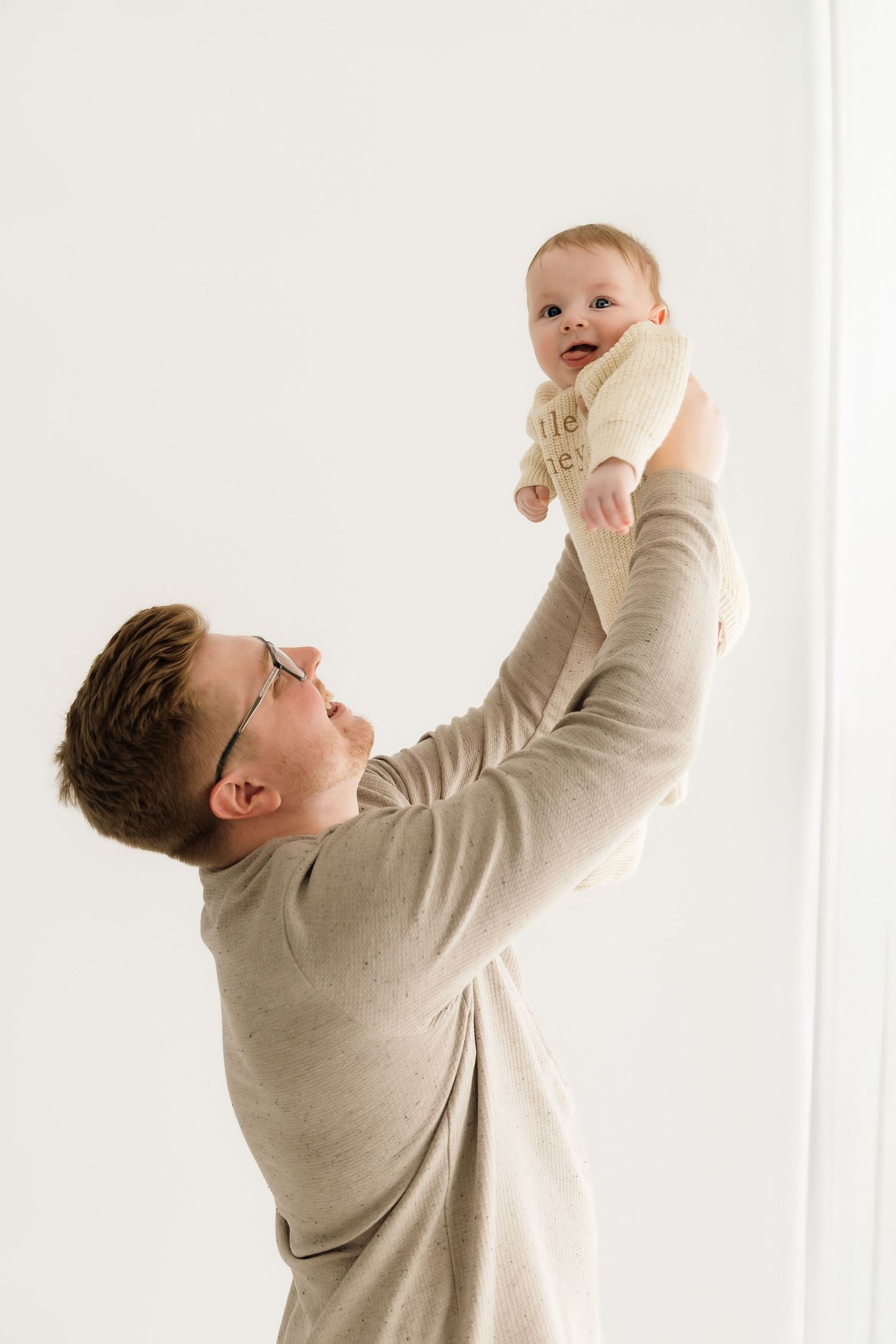 Father in beige sweater joyfully lifting smiling baby in cream knit outfit against white background.