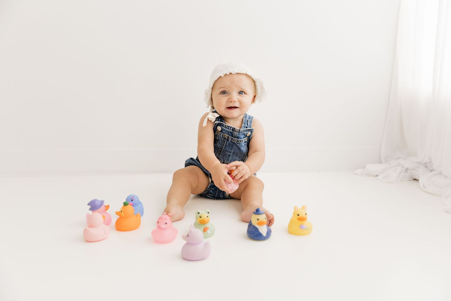 Cute baby girl in denim overalls and white bonnet sitting with colorful rubber ducks on white floor.
