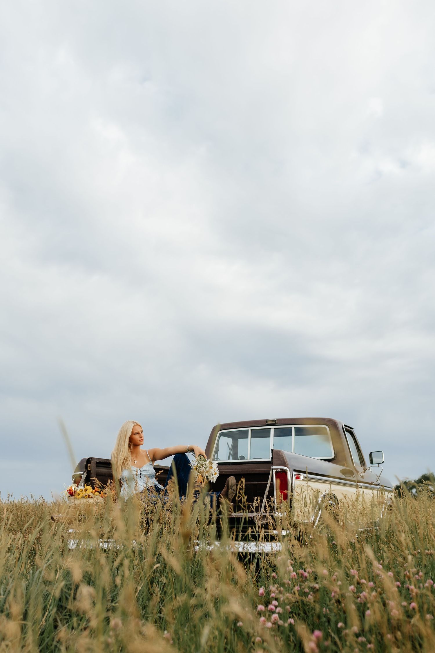 A blonde girl leans against a vintage pickup truck surrounded by tall golden grass under a cloudy sky.