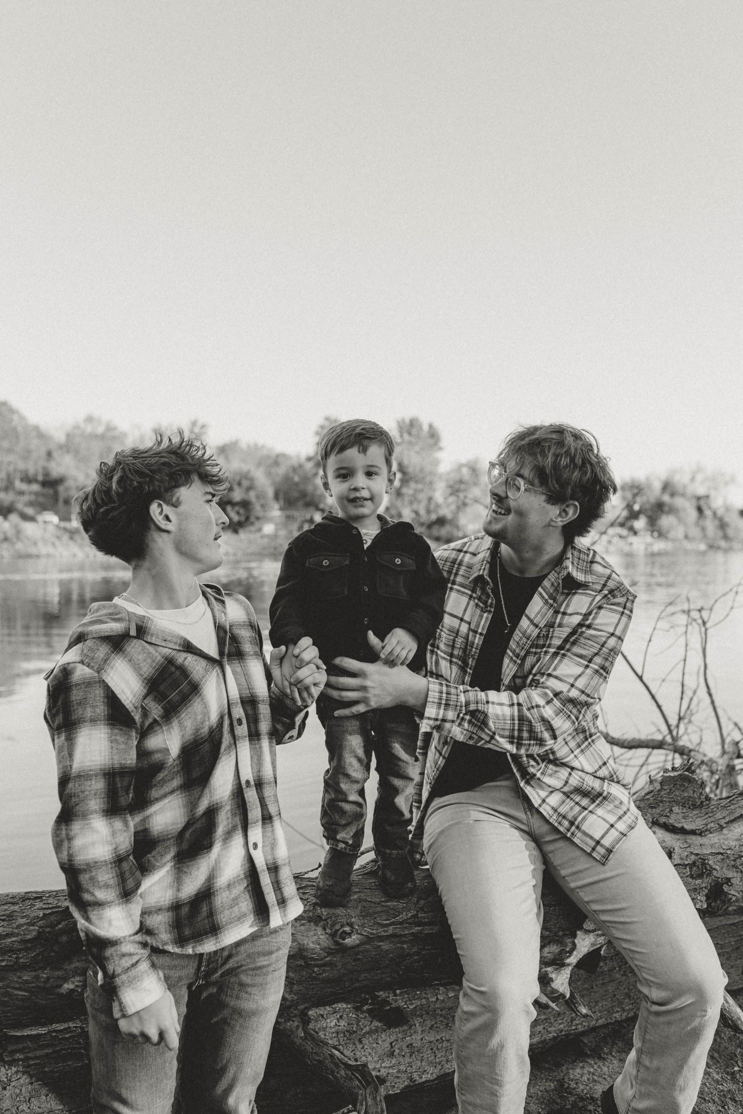 Two teenage boys laugh with toddler standing on log by river in black and white photo.