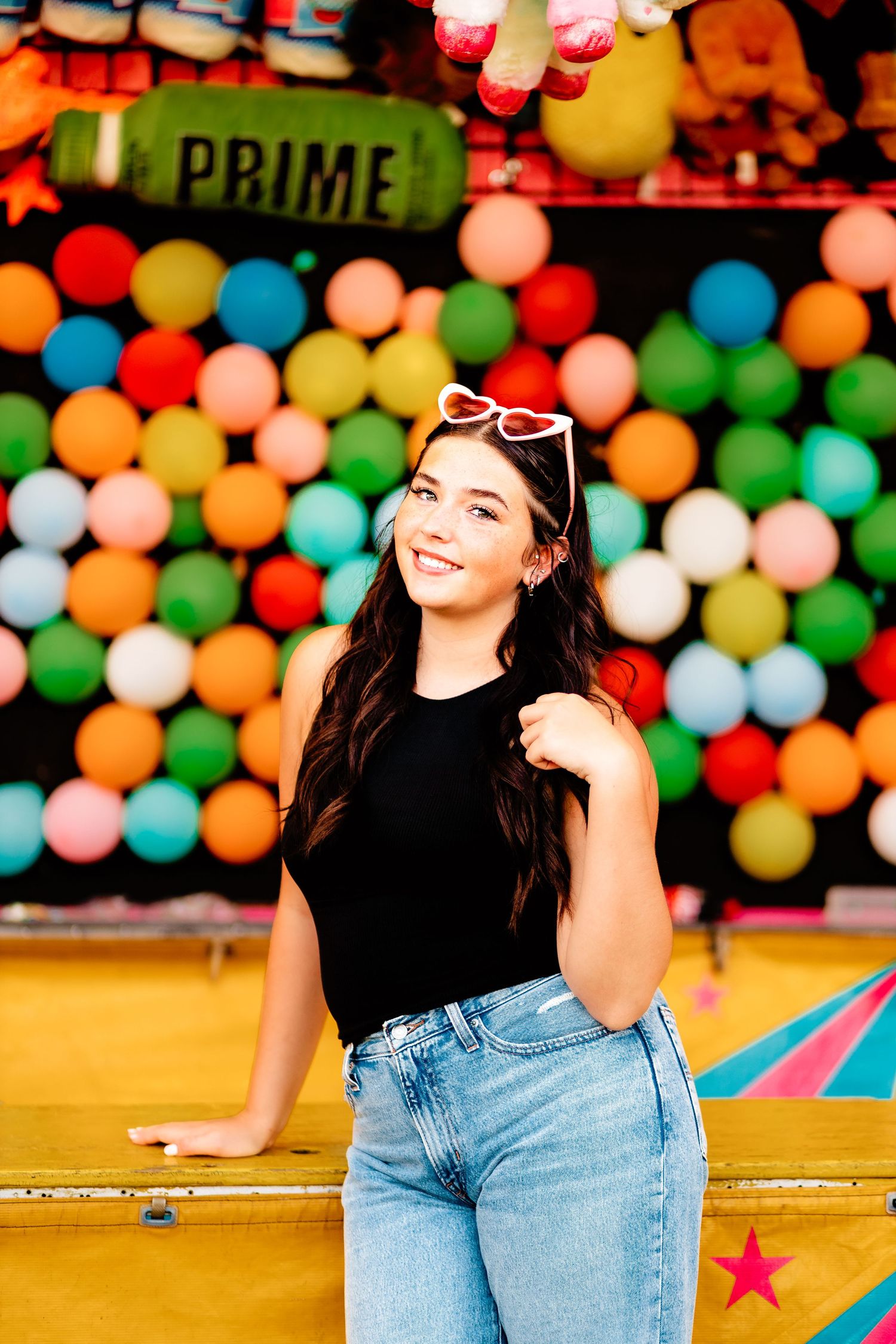 Young woman in black top and jeans smiling at a colorful carnival balloon game booth.