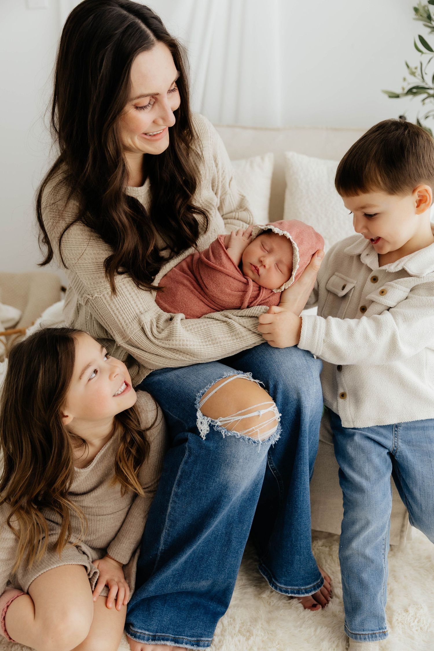 Mother with dark hair sits holding swaddled newborn while two young children gather around admiringly.