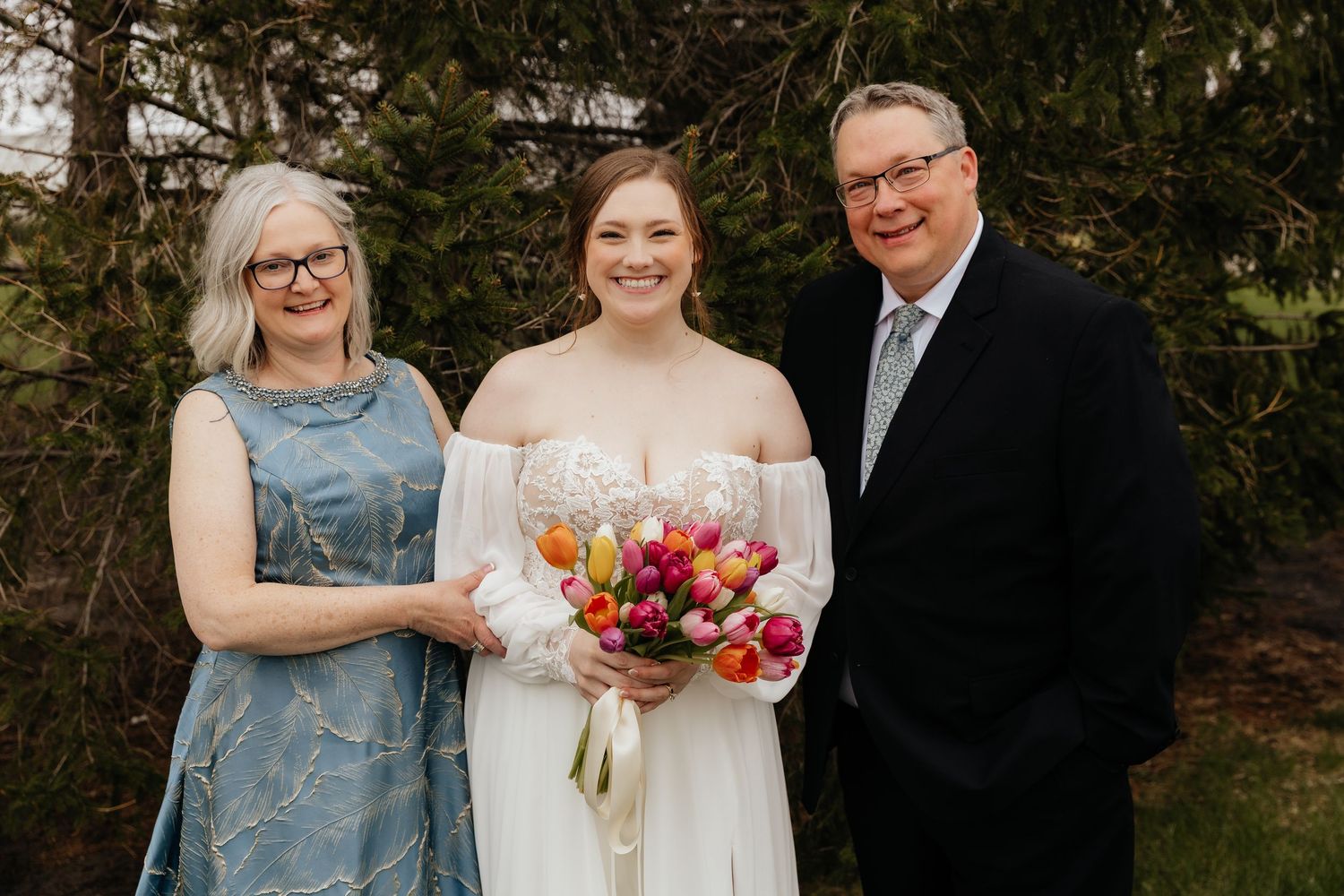 Bride holding colorful tulip bouquet poses with parents in outdoor wedding portrait.
