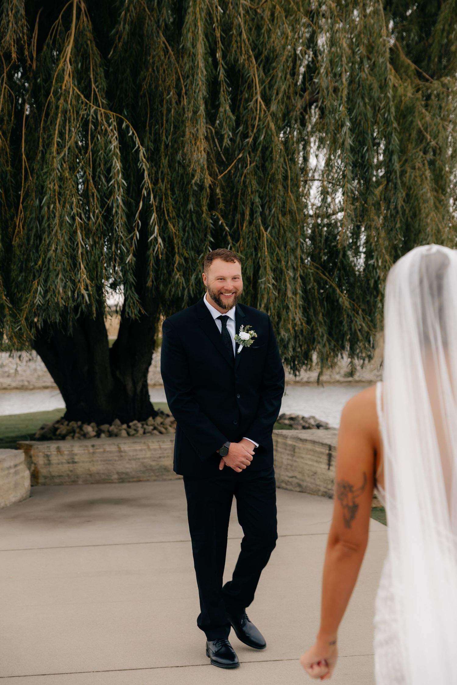 Groom in black suit smiling at bride during first look under weeping willow tree.