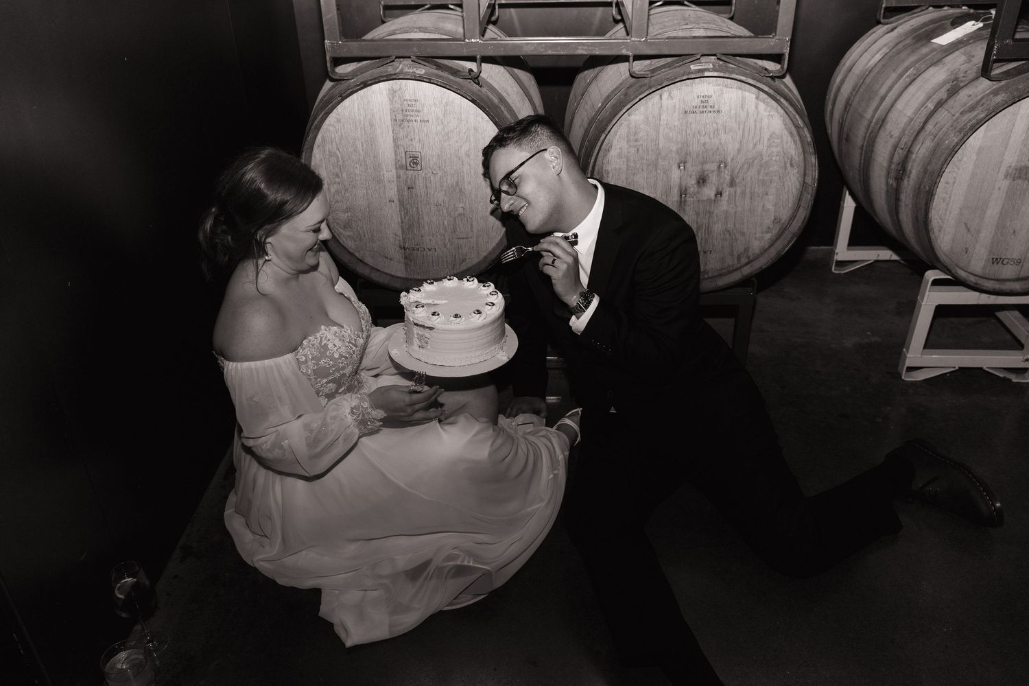 Black and white photo of bride and groom cutting cake near wine barrels.