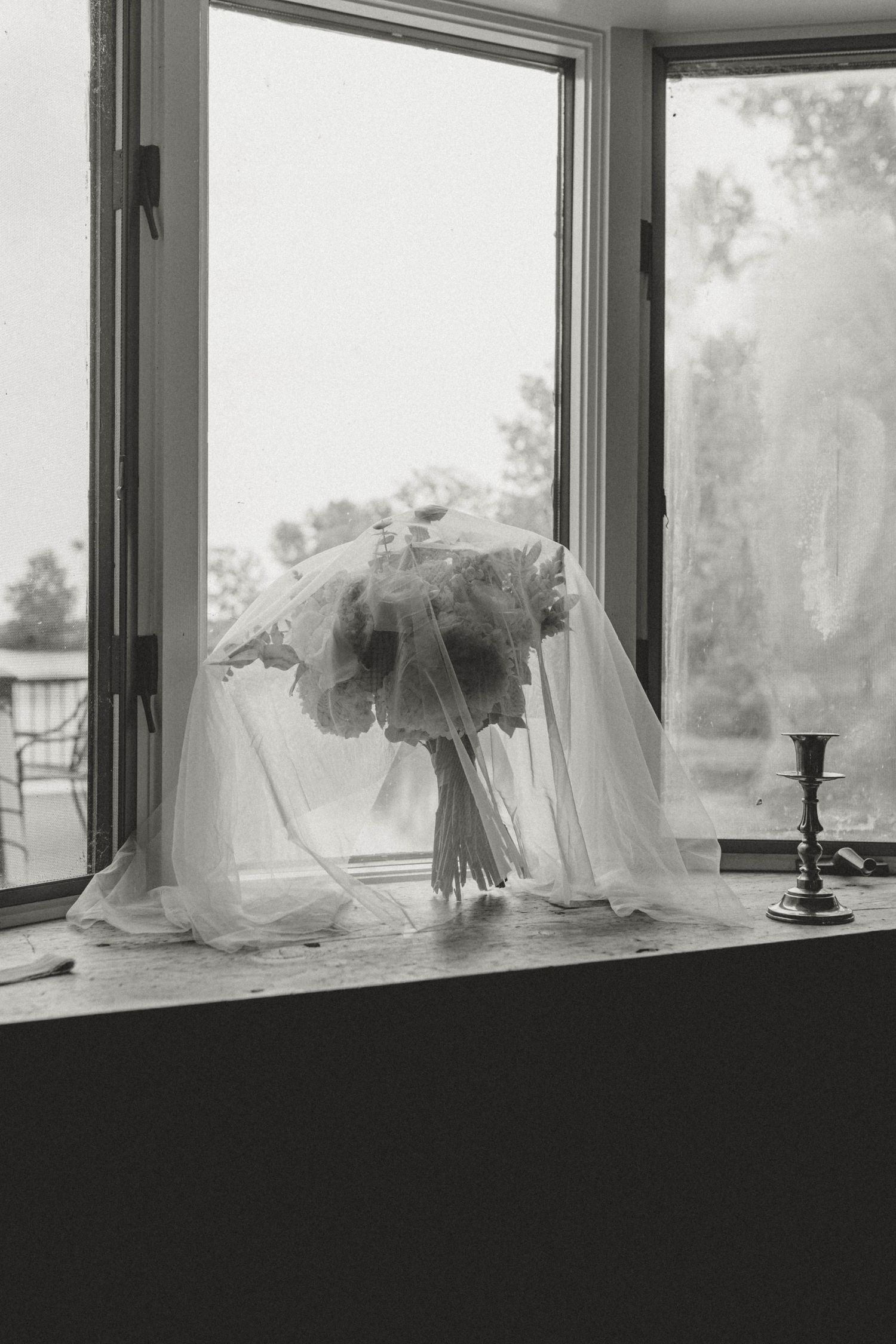 Black and white photo of bridal bouquet draped with veil on window sill with candlestick.
