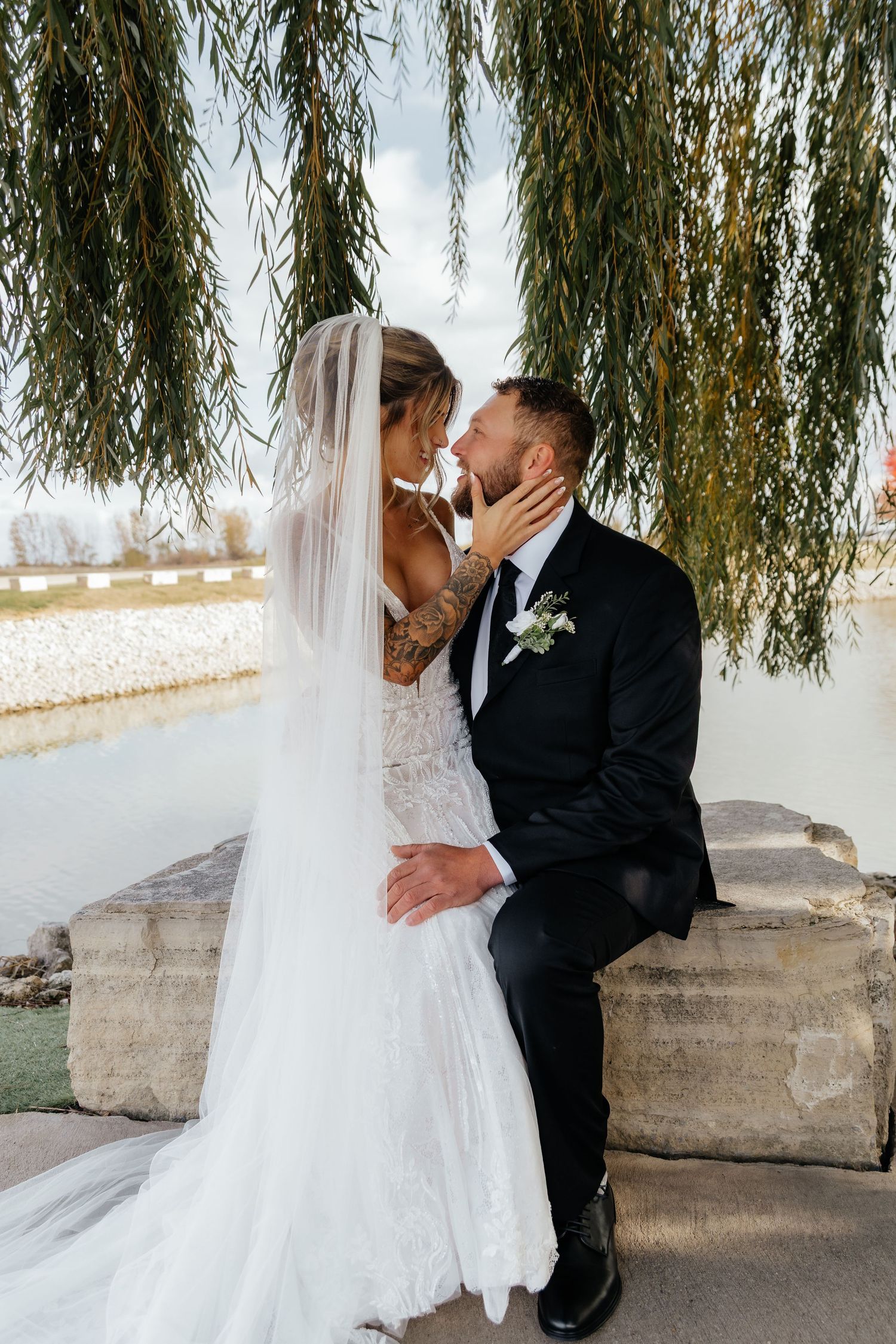 Bride and groom share romantic kiss near waterfront under weeping willow, groom seated on stone wall.