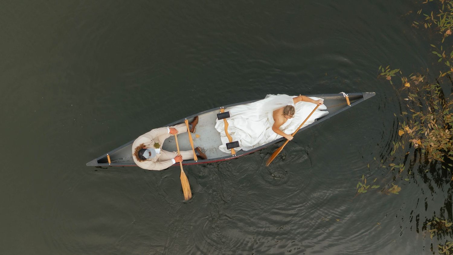 Aerial view of a bride and groom paddling a canoe on a calm river.