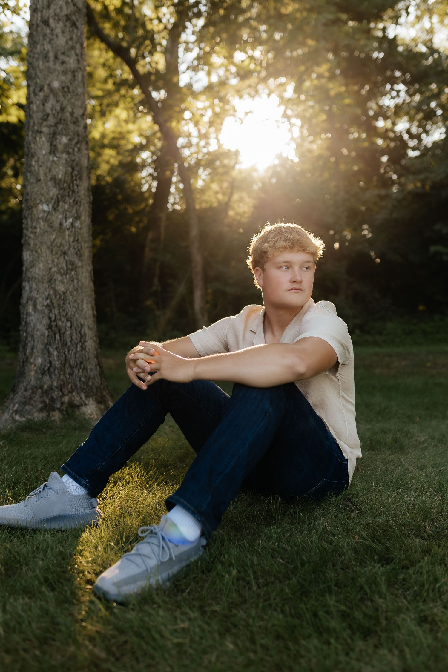 Young man in a beige shirt and jeans sitting on grass leaning against a tree at golden hour.