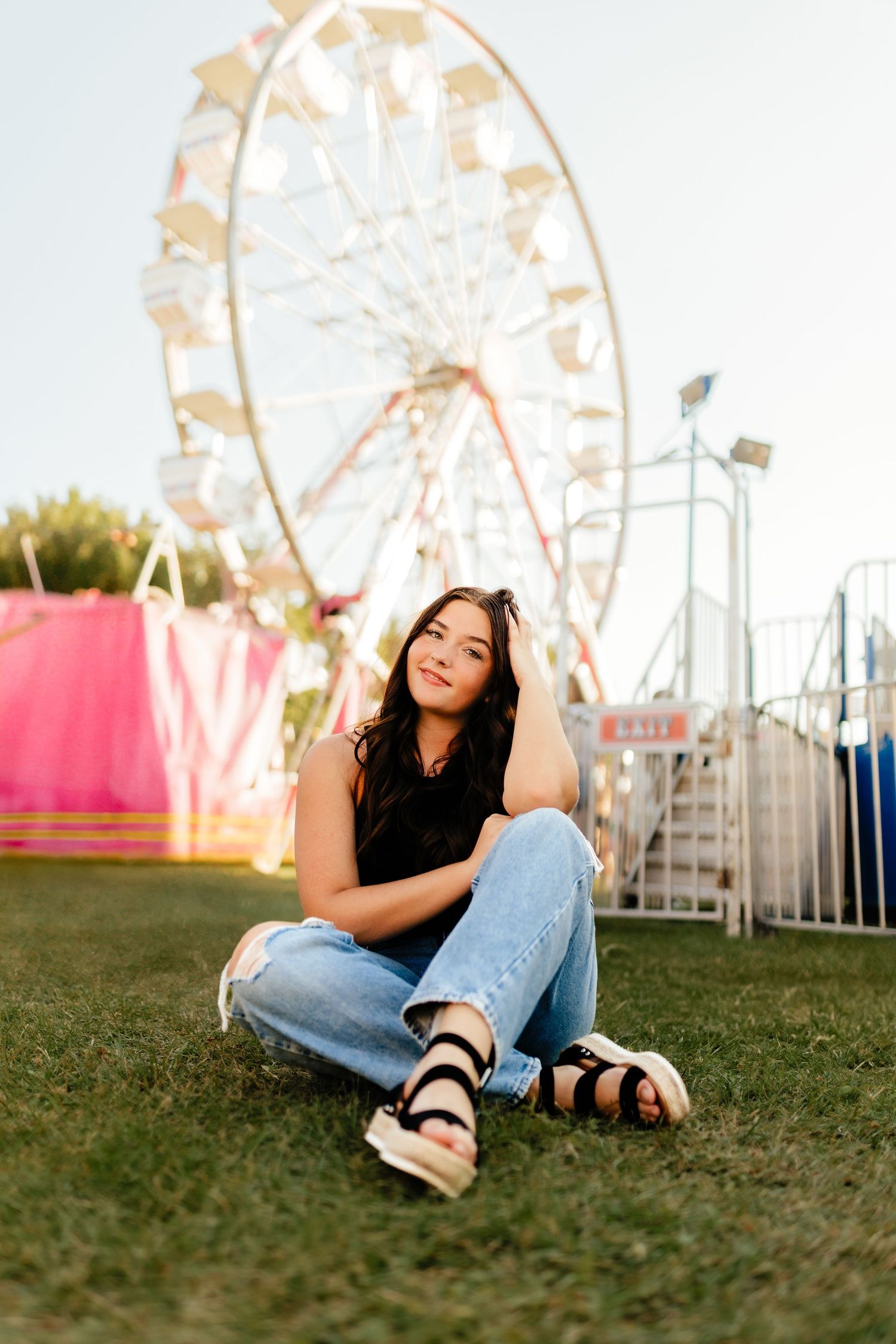 Young brunette woman sitting on grass in front of a ferris wheel at a sunny outdoor fairground.