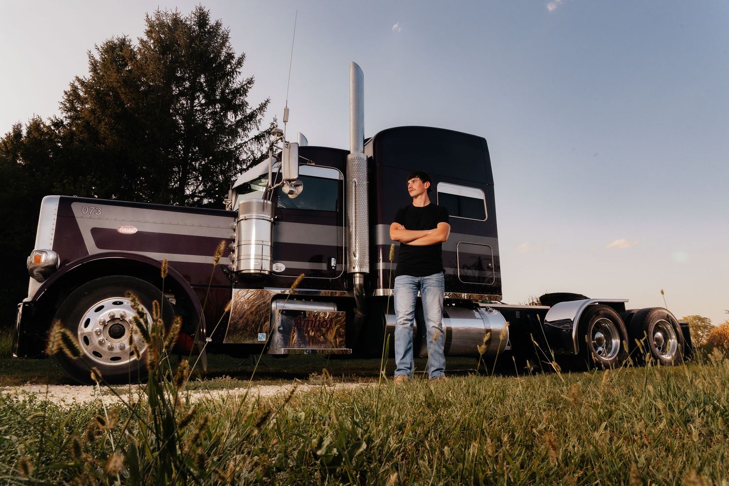 Man in black shirt and jeans stands with arms crossed beside a large black semi truck outdoors.