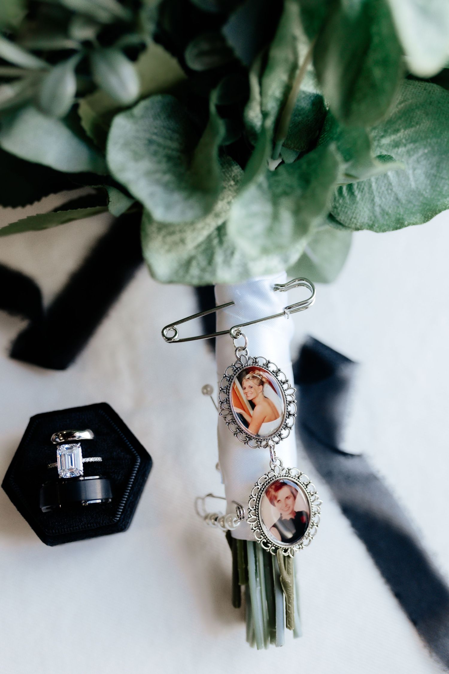 Wedding bouquet with memorial photo charms pinned to white ribbon wrap and diamond ring in black box.