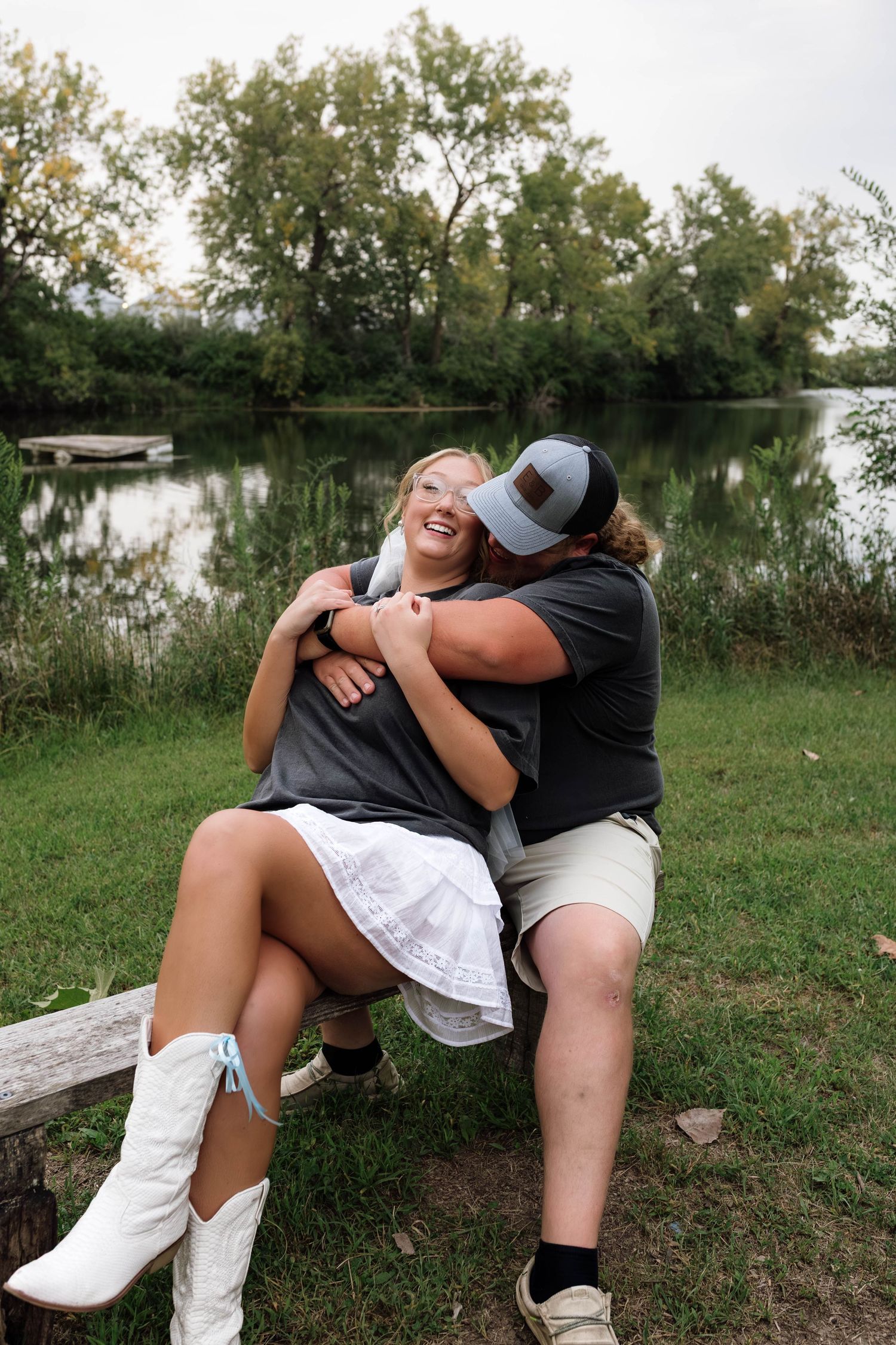 Happy couple sitting by a lakeside, embracing and laughing in casual wedding attire.