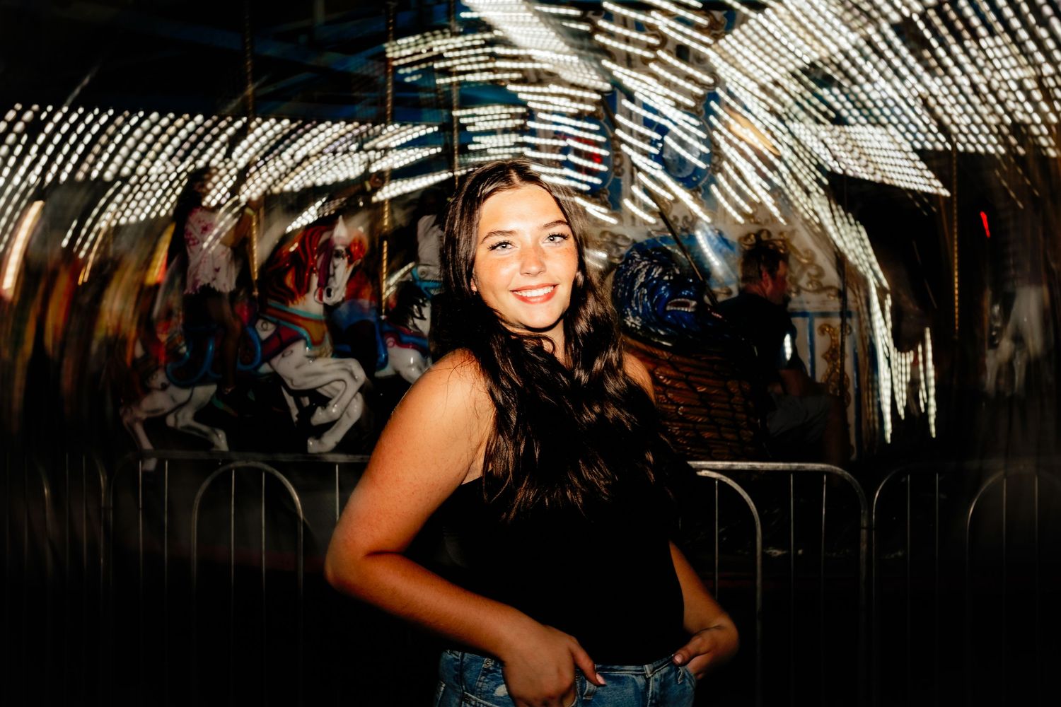Young woman smiling in front of a spinning carousel with bokeh lights at night fair.
