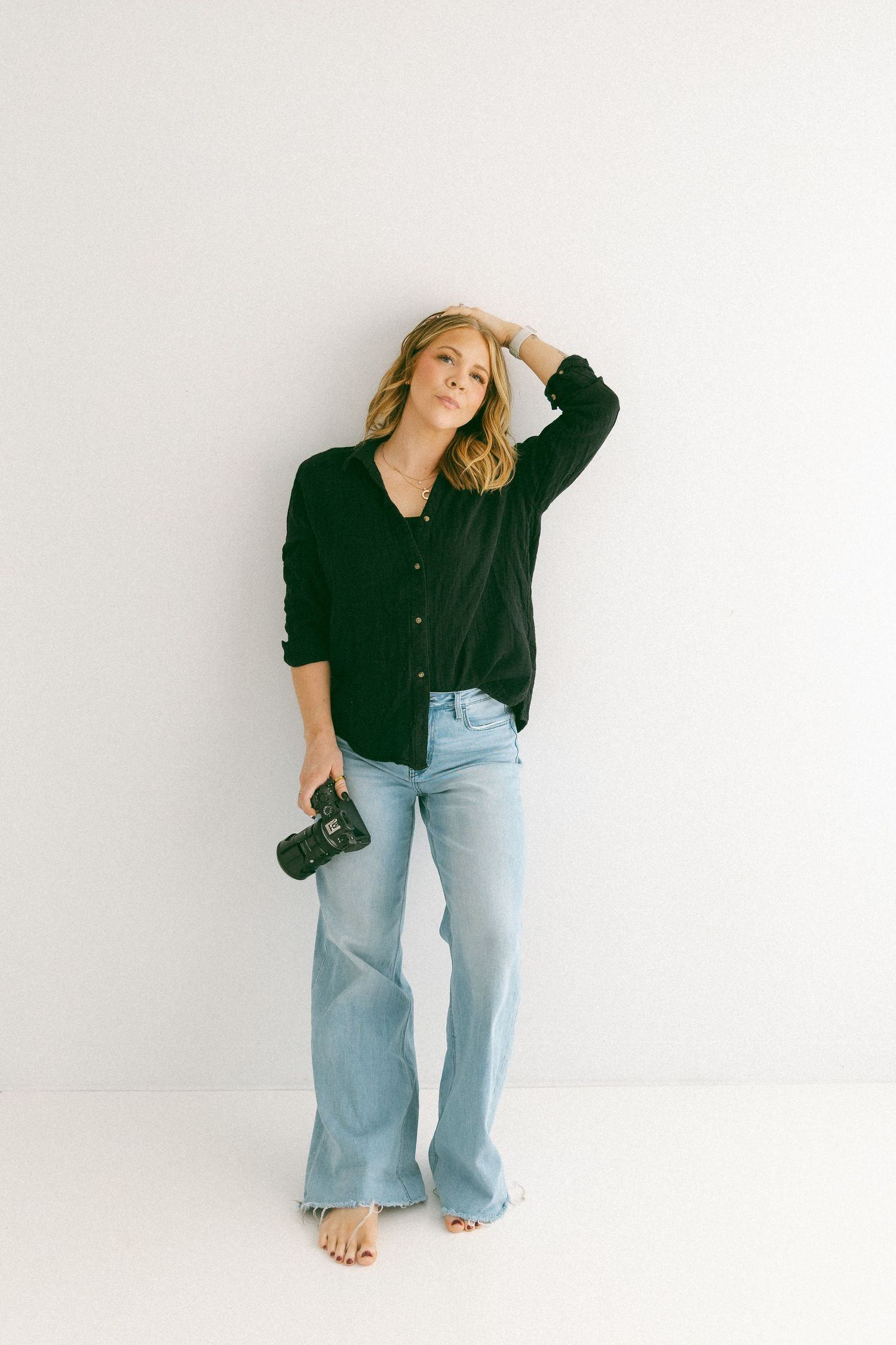 Blonde female photographer in black shirt and jeans holding a camera against a white studio wall.