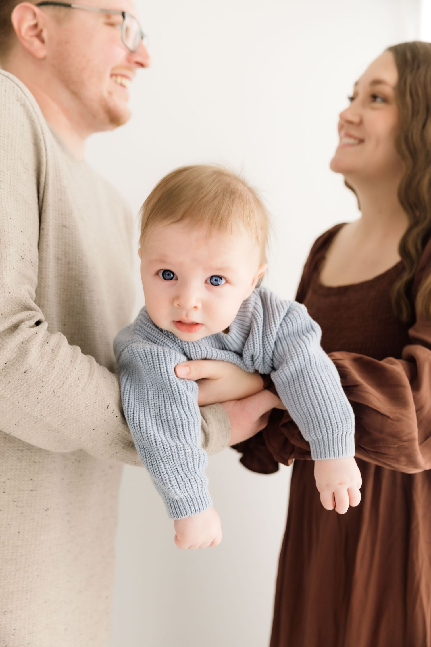 Smiling parents holding their blue-eyed baby in a light blue knit sweater.
