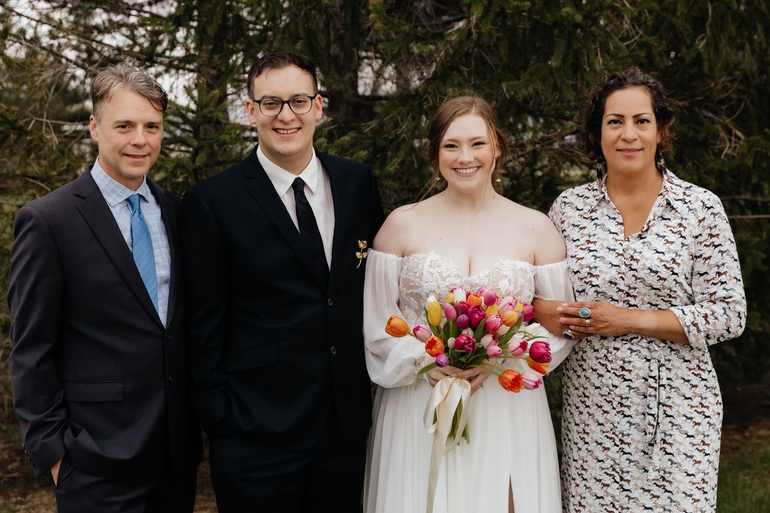 Bride holding tulip bouquet poses with groom and two guests outdoors in a garden setting on wedding day.