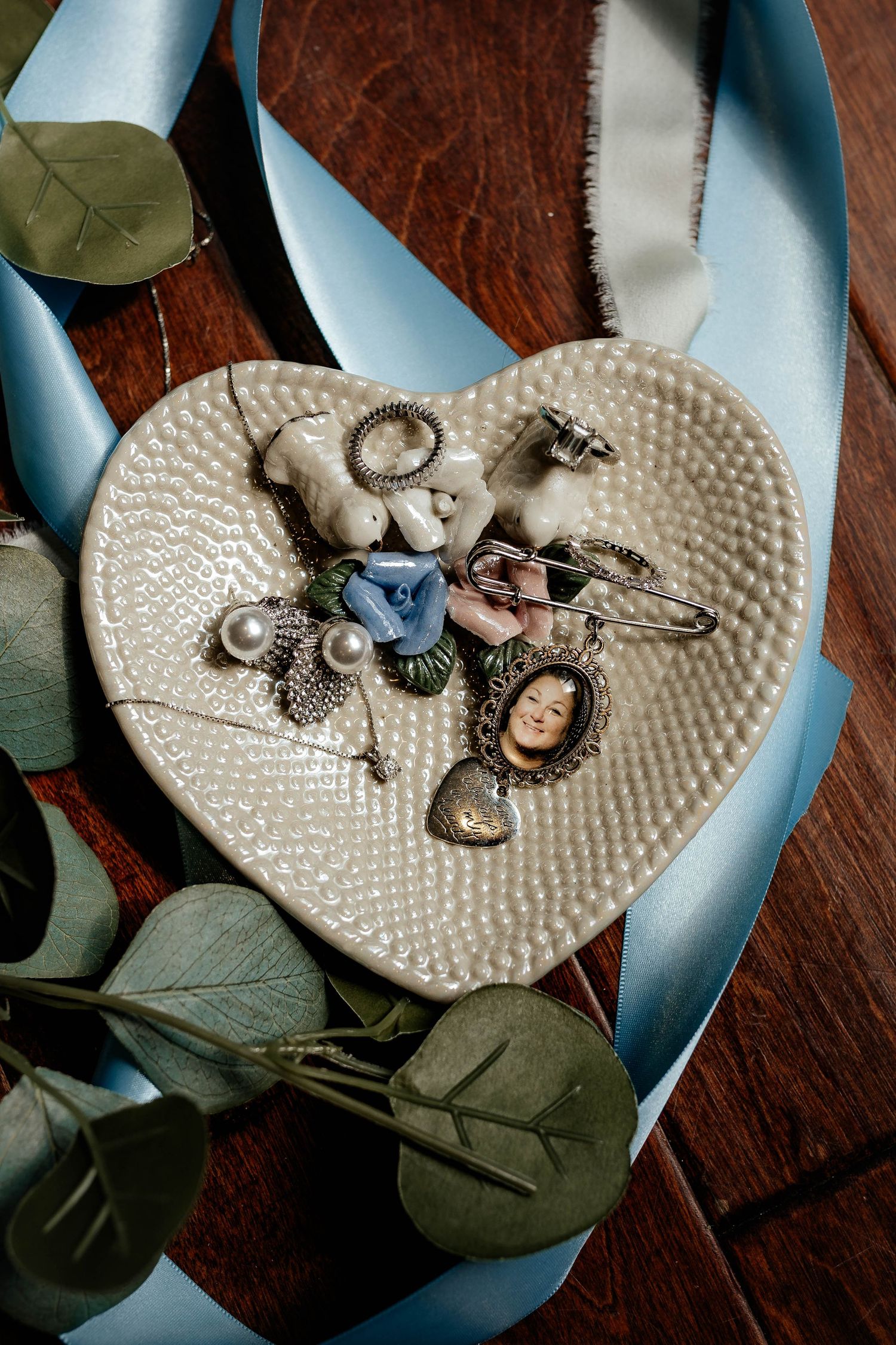 Heart-shaped ceramic dish with vintage brooches, pearls, and a cameo surrounded by eucalyptus and blue ribbon.